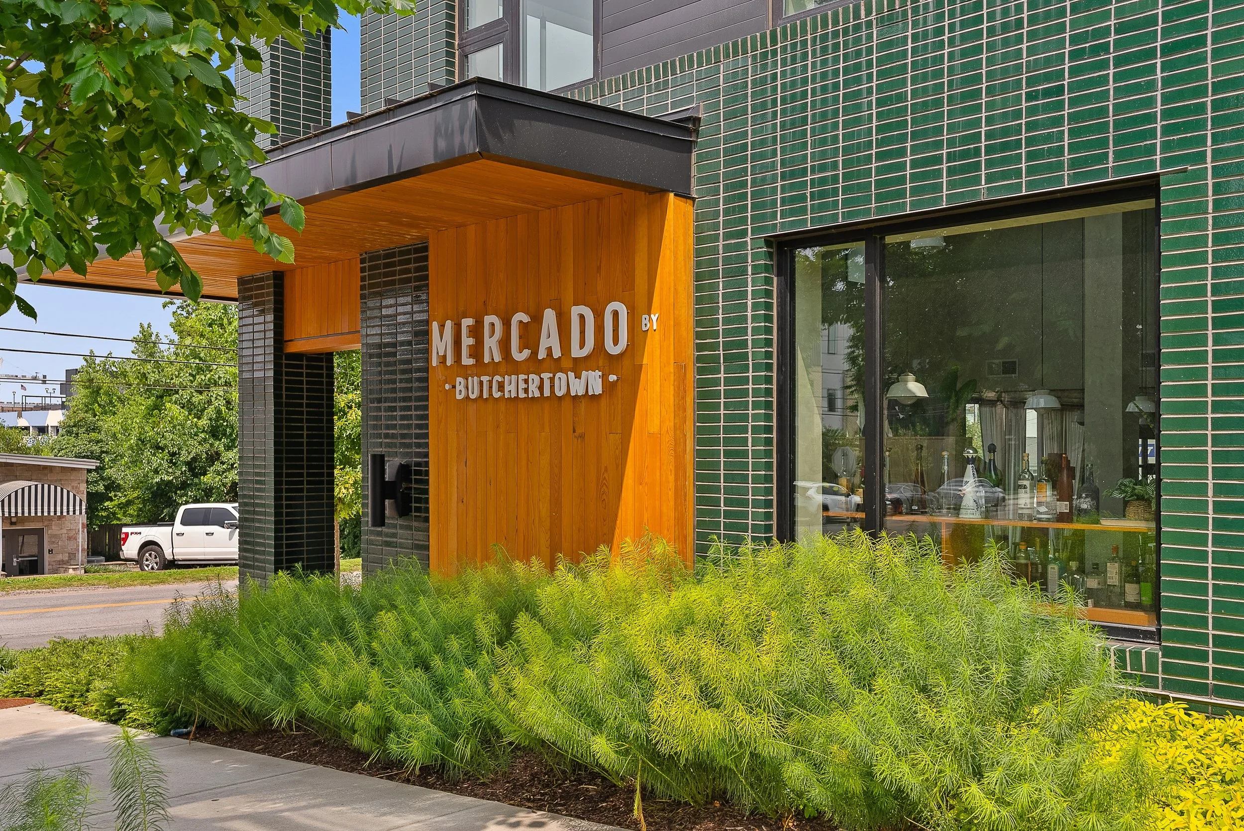 Exterior of Mercado by Butchertown, a modern building with green brick walls, a wooden entrance sign, and lush green bushes in front.