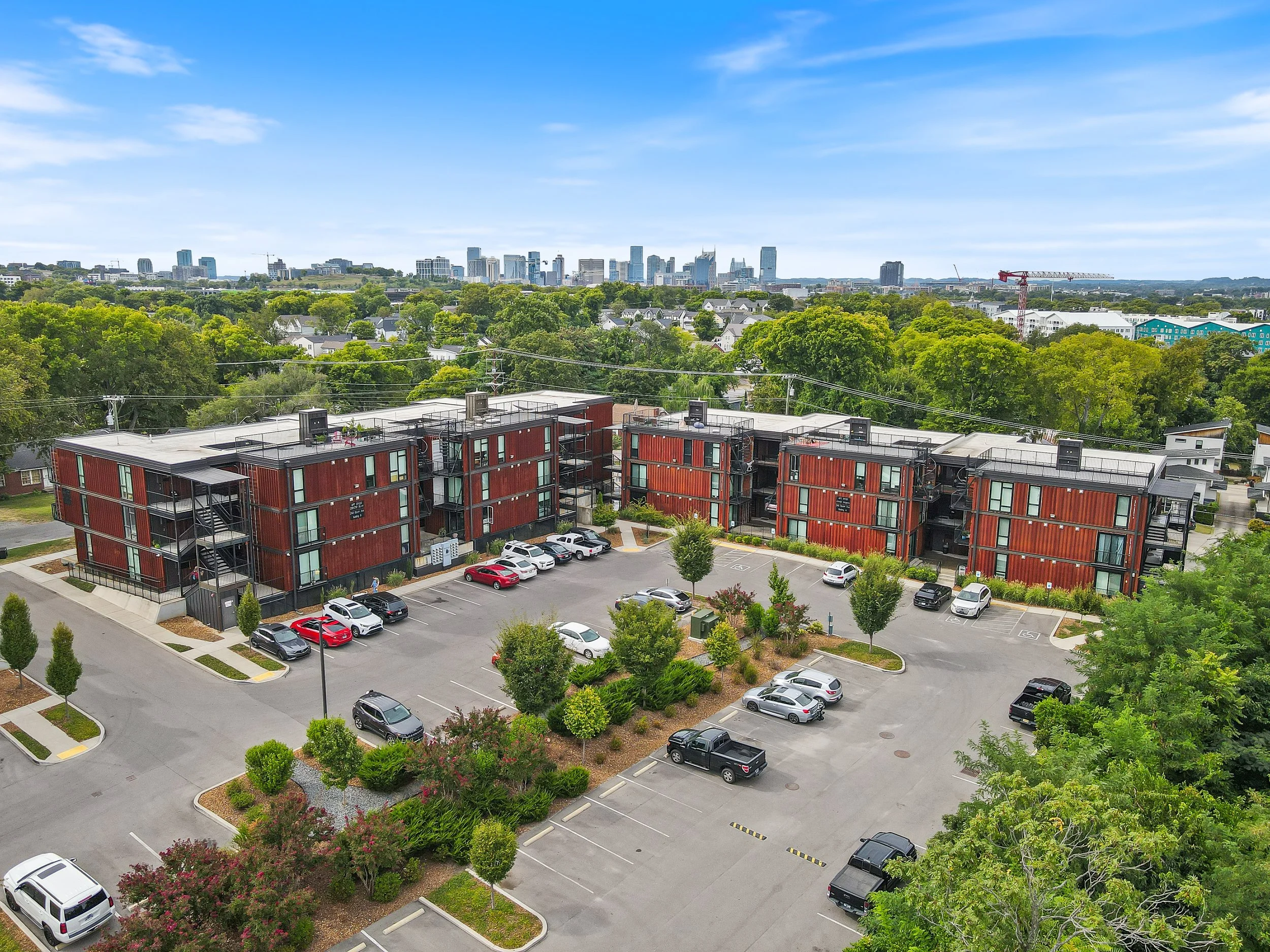 Aerial view of a modern apartment complex with a parking lot, surrounded by trees and greenery, with a city skyline in the background.