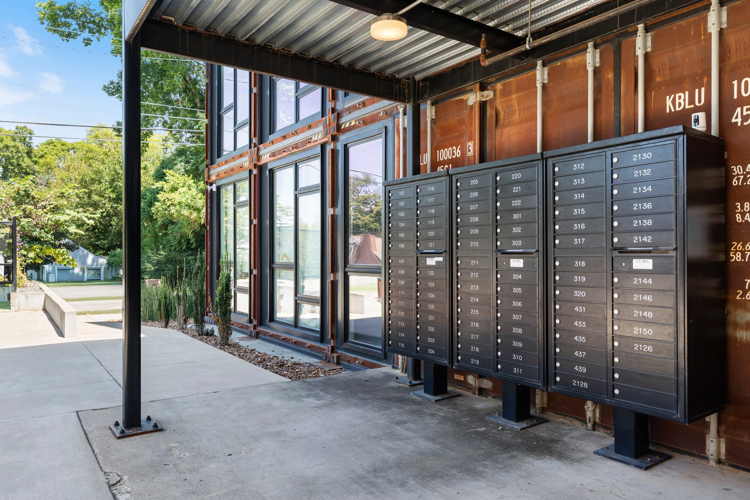 Mailboxes in a modern building with large windows and a concrete sidewalk outside, surrounded by trees.