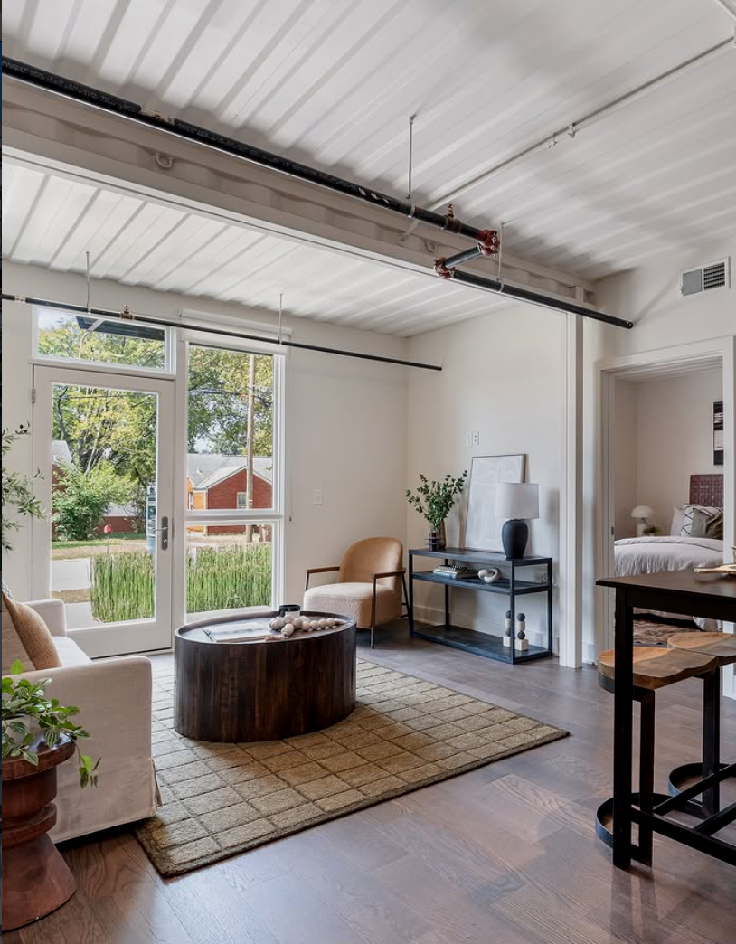 Modern living room with large glass door leading outside, beige sofa, single beige armchair, round dark wood coffee table, area rug, side table with lamp and plant, and hardwood floors.