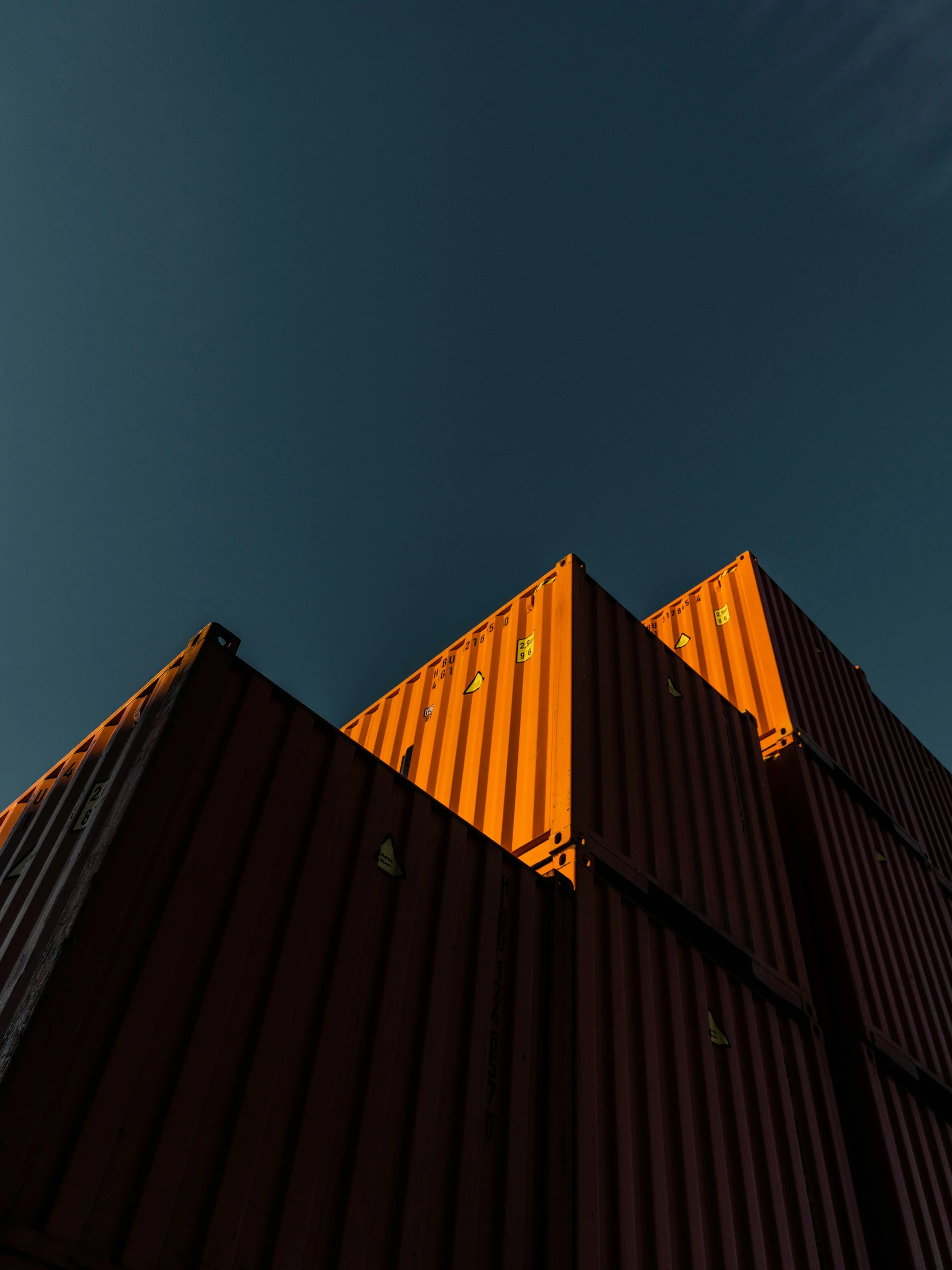 Stacked orange and gray shipping containers viewed from below against a dark blue sky.