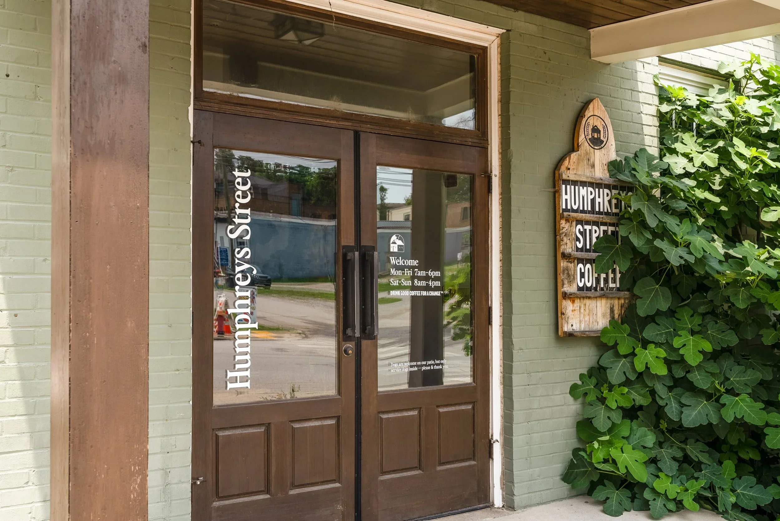 Green brick building entrance with wooden door and sign for Humphrey Street Coffee, featuring a leafy plant nearby.