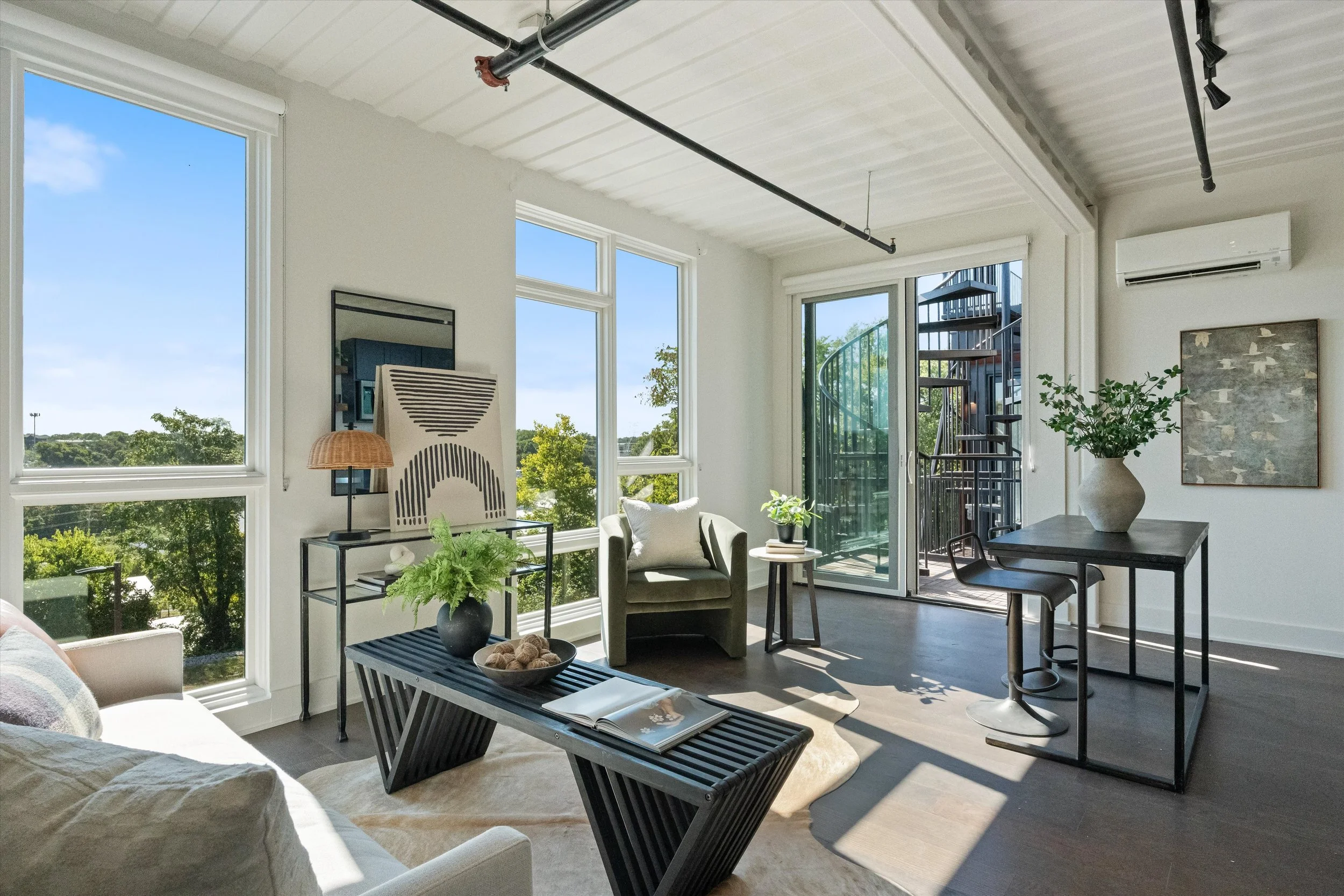 Bright living room with large windows, a white sofa, black coffee table, beige armchair, side table with plant, black dining table with white vase, and spiral staircase outside sliding glass door.