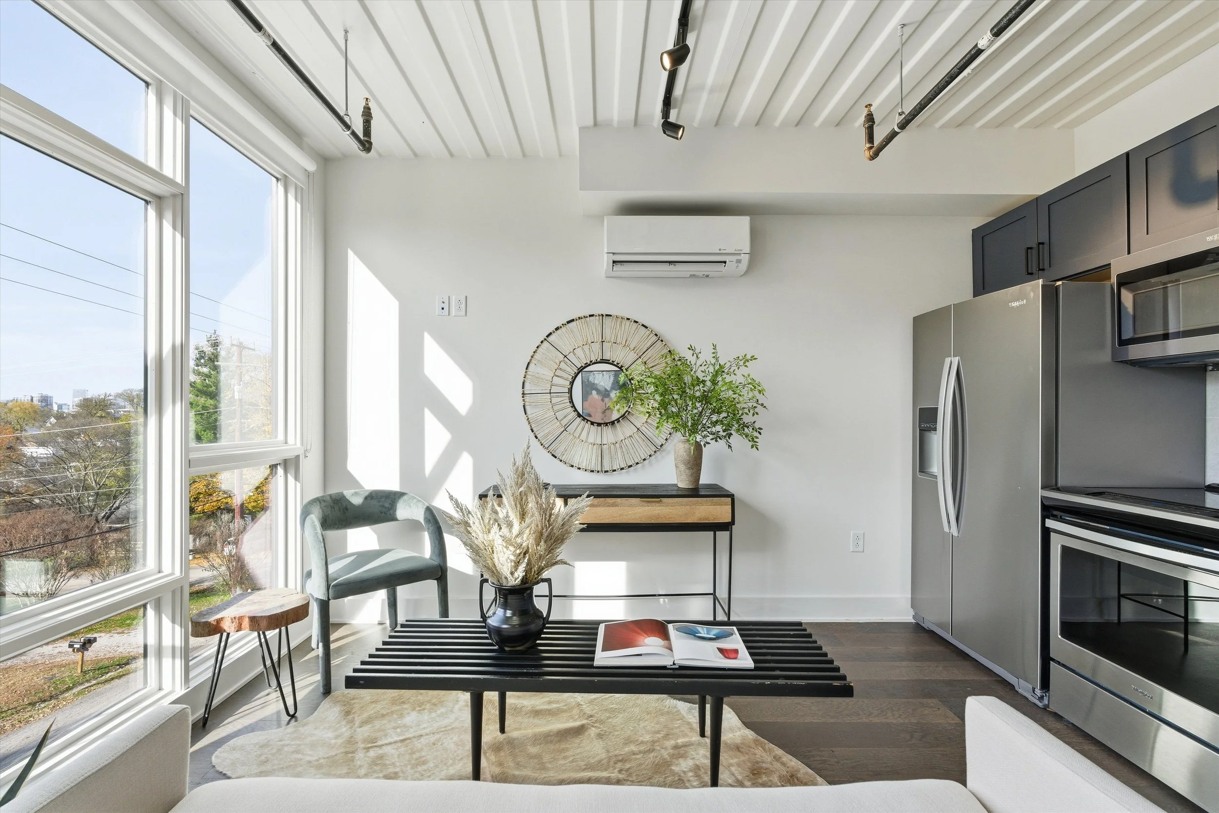 Bright living room with large windows, modern kitchen appliances, a black coffee table with a vase of dried flowers and an open magazine, a chair, a small side table, and decorative wall mirror.