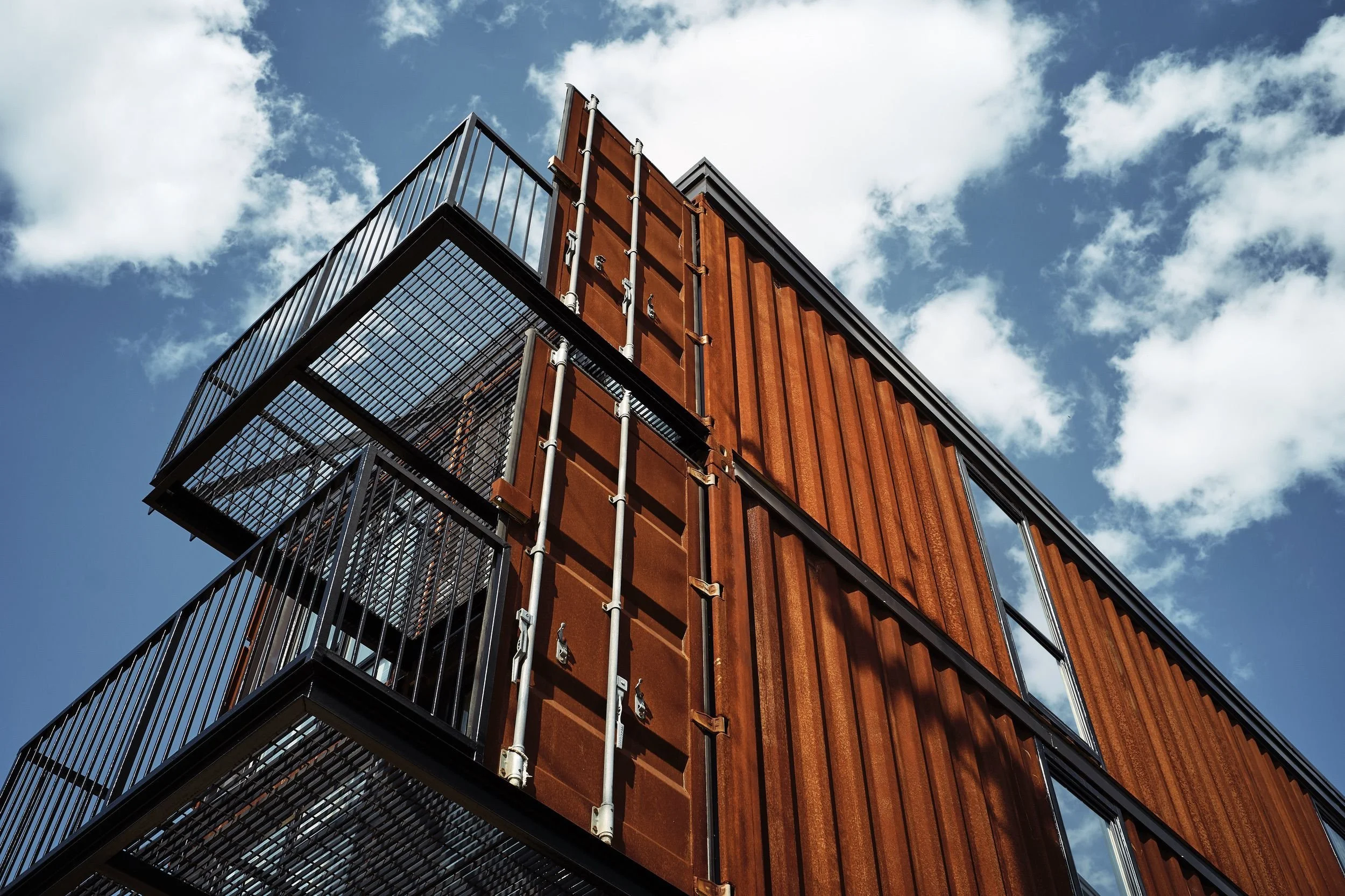 Modern building with rust-colored metal siding and black metal balconies, against a bright blue sky with scattered clouds.