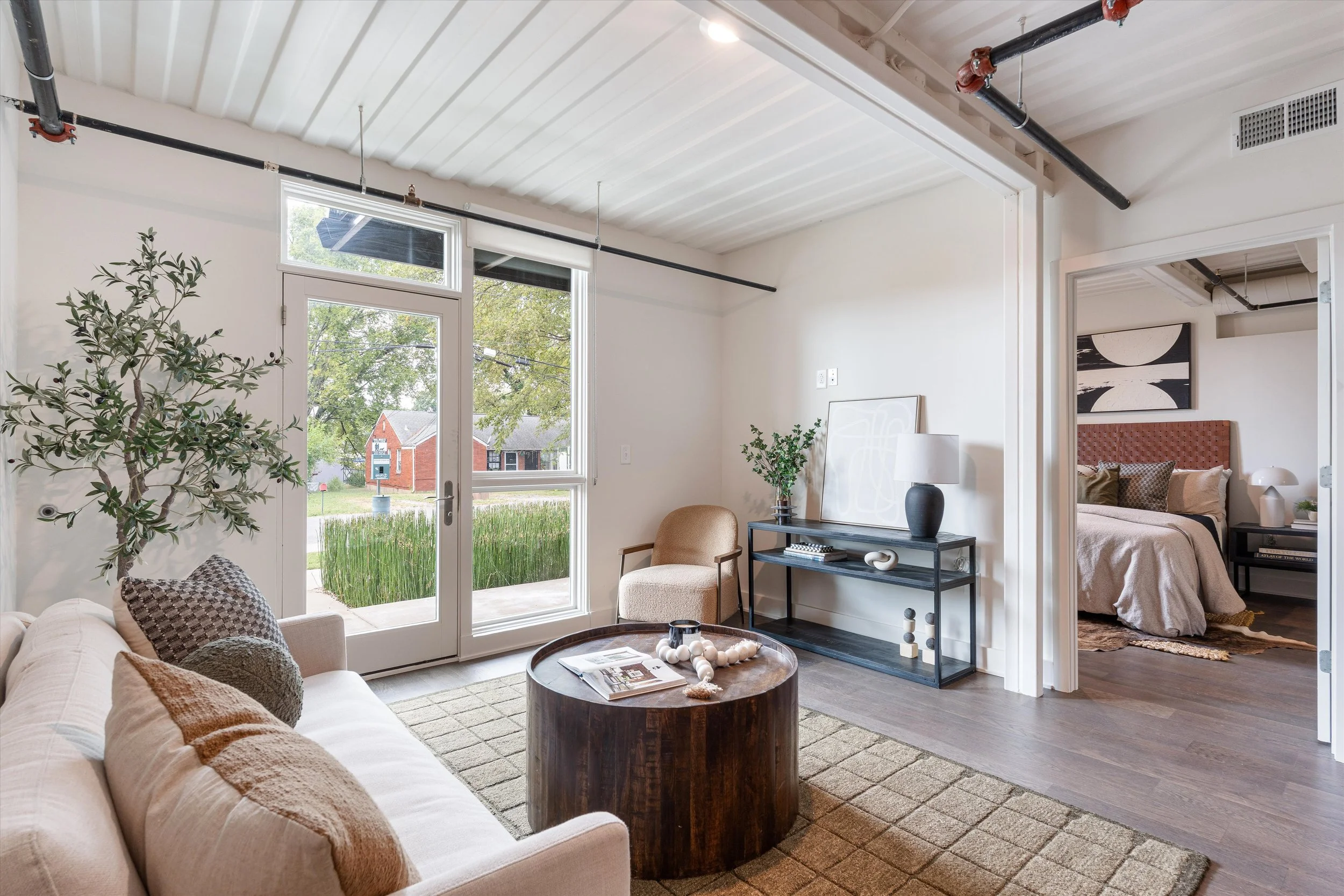 Living room with beige sofa, coffee table, green plant, and large windows showing outdoor view. Bedroom with bed and art over head in the background.