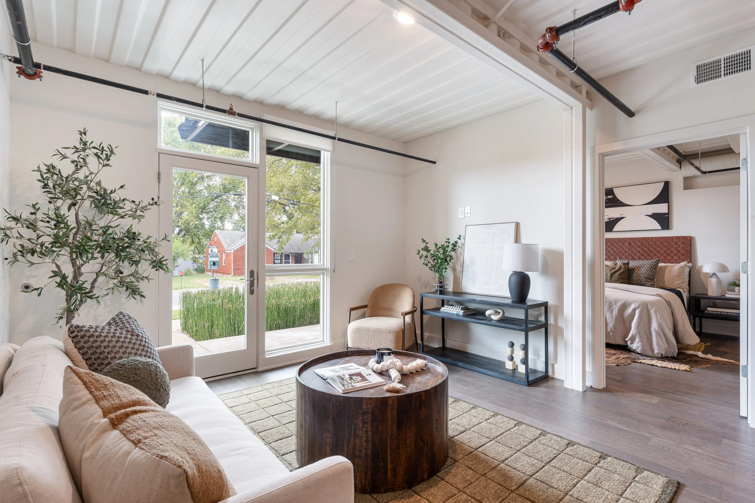 Living room with large window and glass door, beige sofa with pillows, round wooden coffee table, armchair, black console table with decorative items, and view into a bedroom with bed and artwork on the wall.