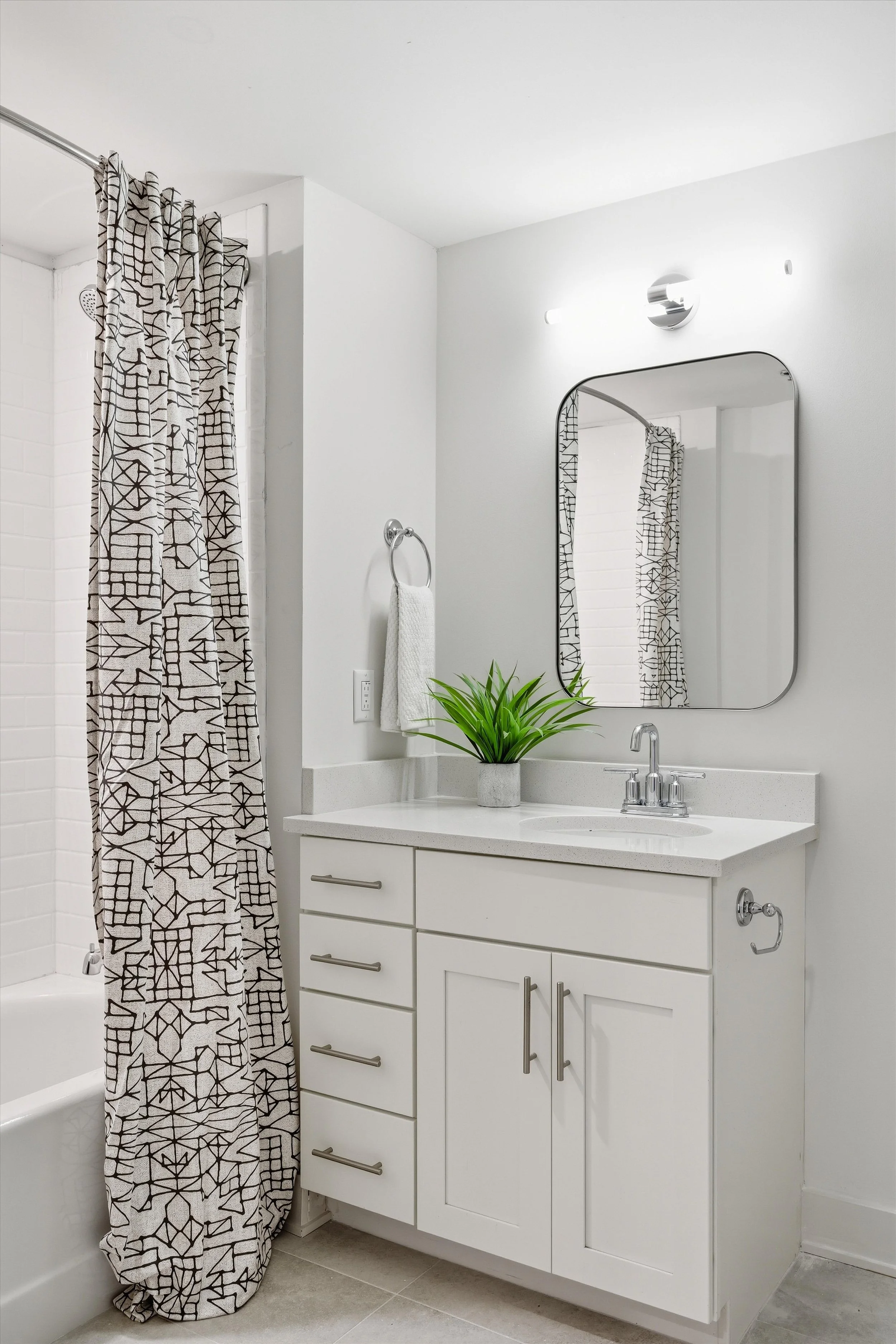 Modern bathroom with a white vanity, a large mirror, a potted green plant, a towel on a ring, and a patterned shower curtain.