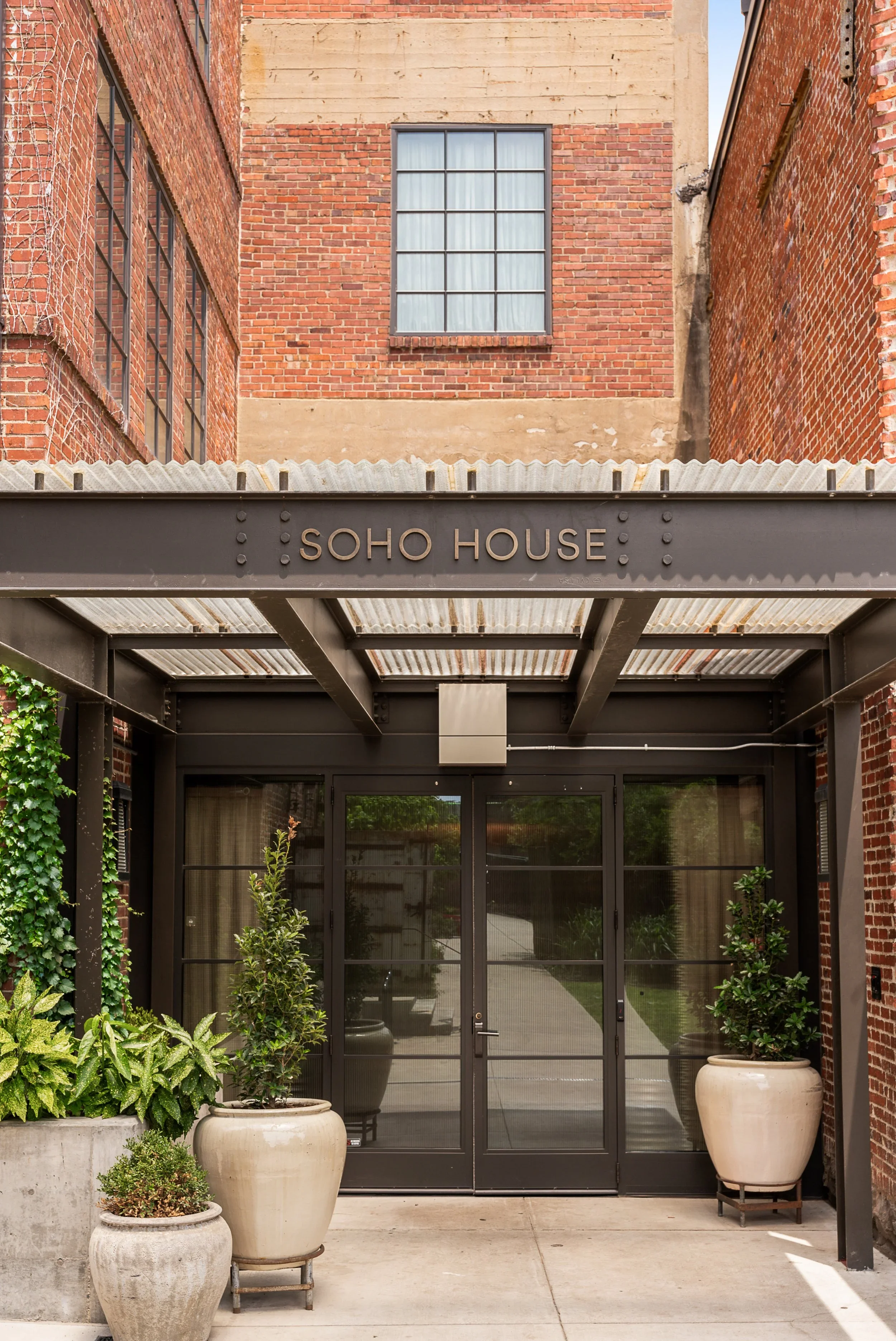 Entrance to Soho House with black metal frame glass doors, potted plants, and a sign above the entrance that reads 'SOHO HOUSE'.