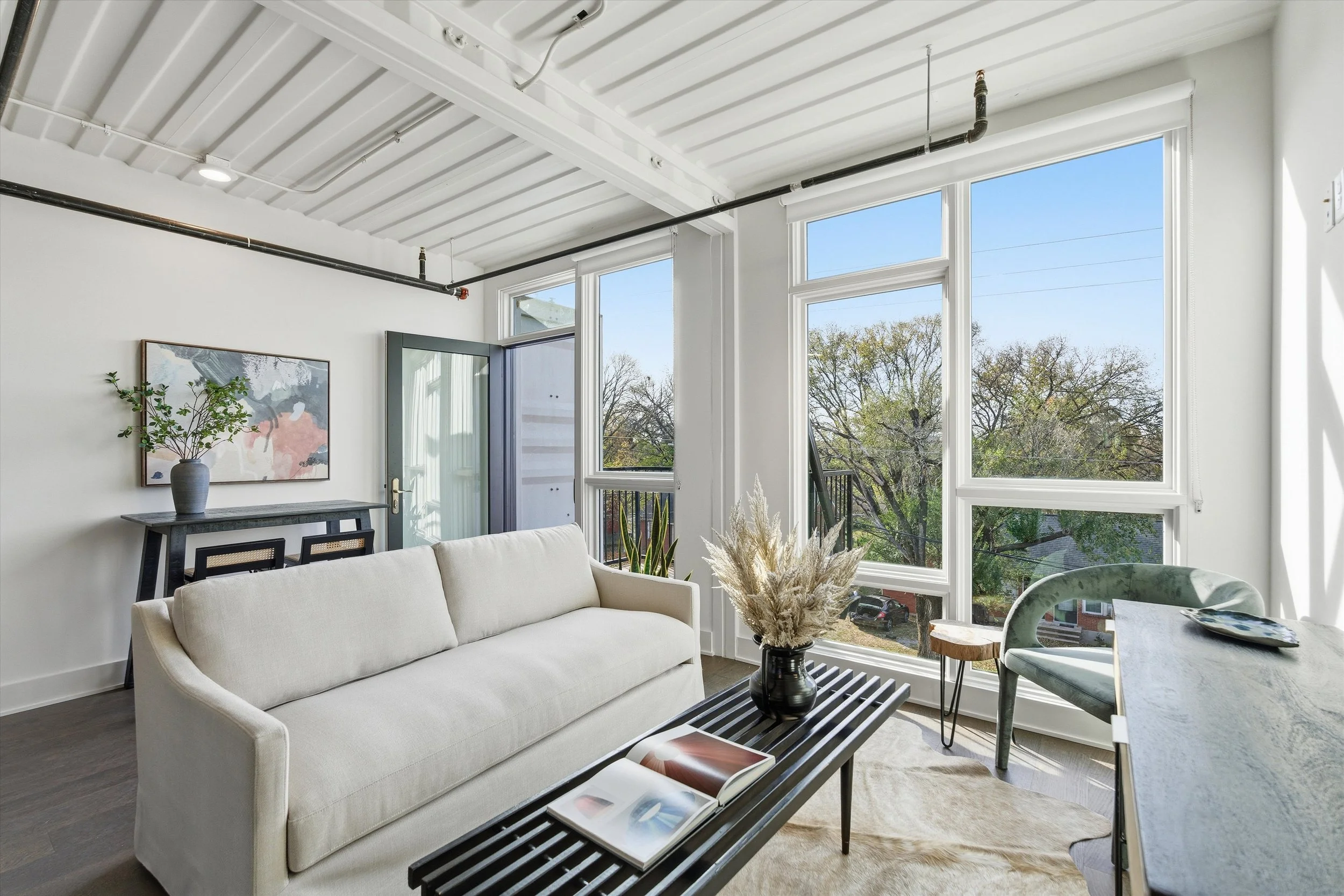 Bright living room with large windows, white sofa, black coffee table with magazine and vase of dried flowers, green chair at a desk, and artwork on the wall.
