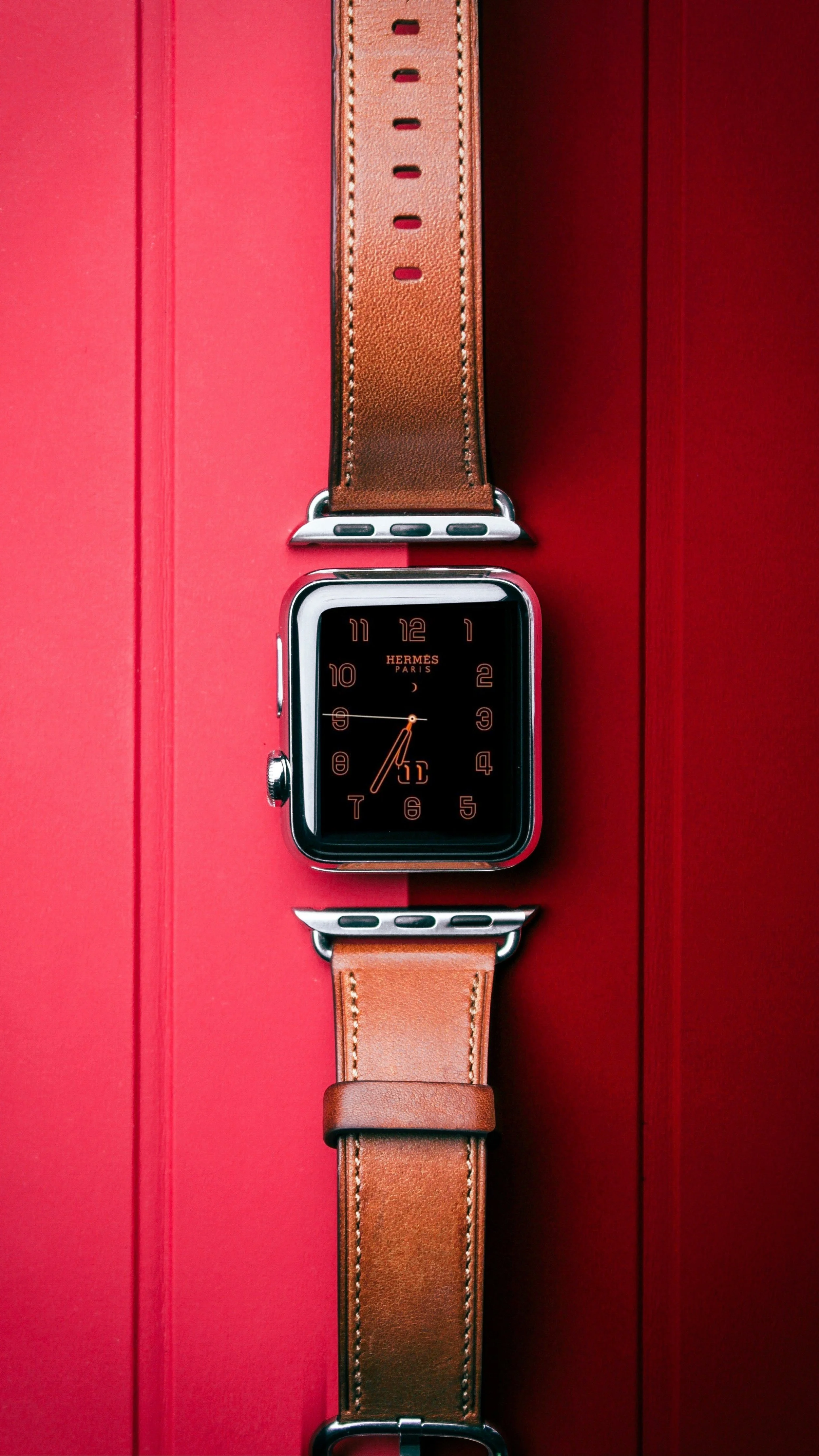 An Hermes smartwatch with a brown leather band displaying the time, placed on a red surface.