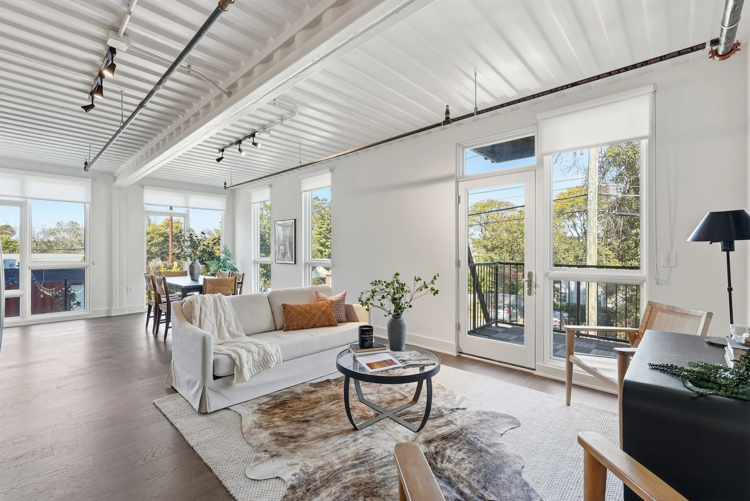 Bright living room with large windows, white walls, wood floors, a white sofa with pillows, a round coffee table, and a dining area in the background.