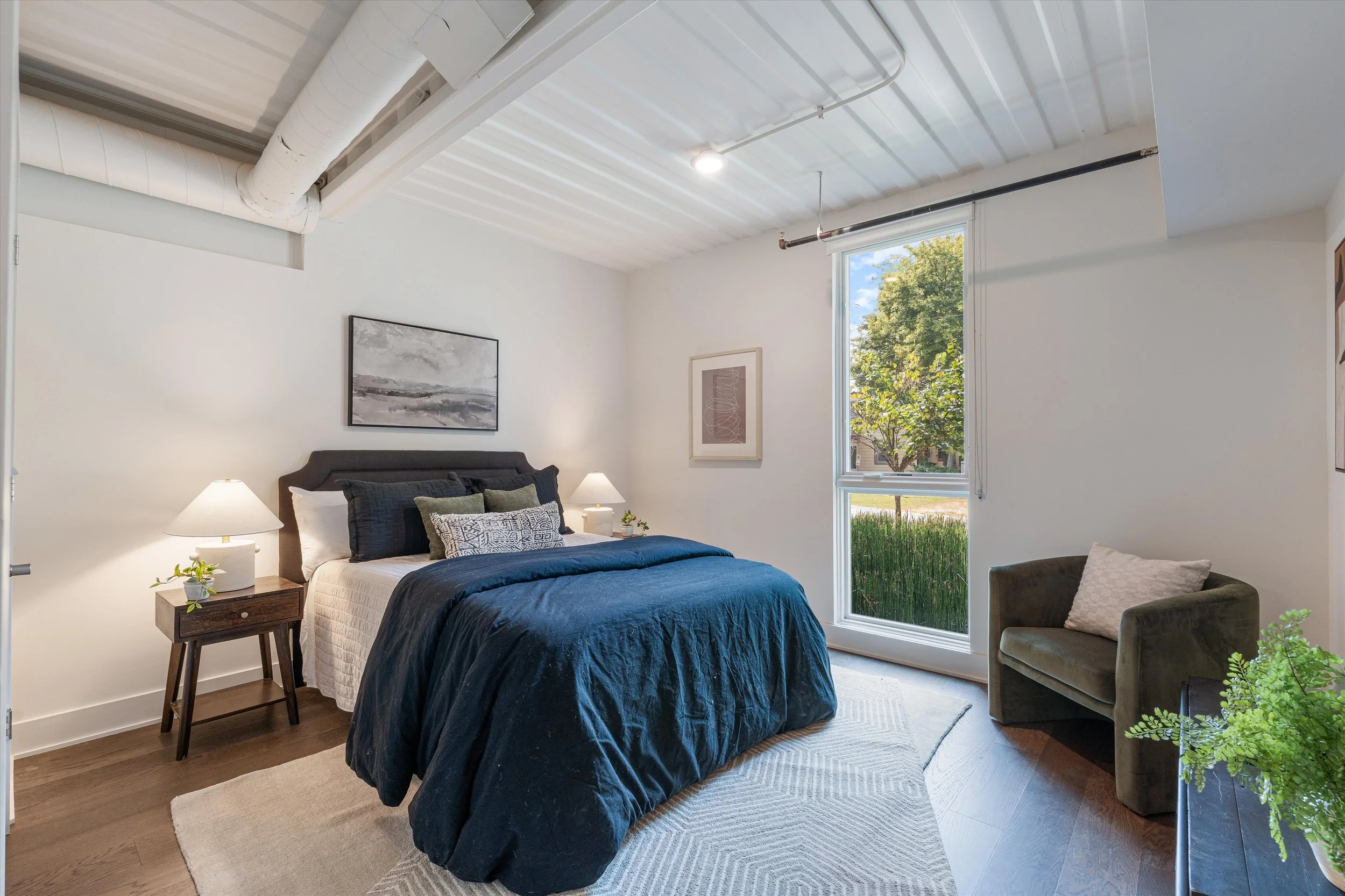 A bedroom with a bed, side tables with lamps, a large window showing trees outside, a green armchair with a pillow, and artwork on the white walls.