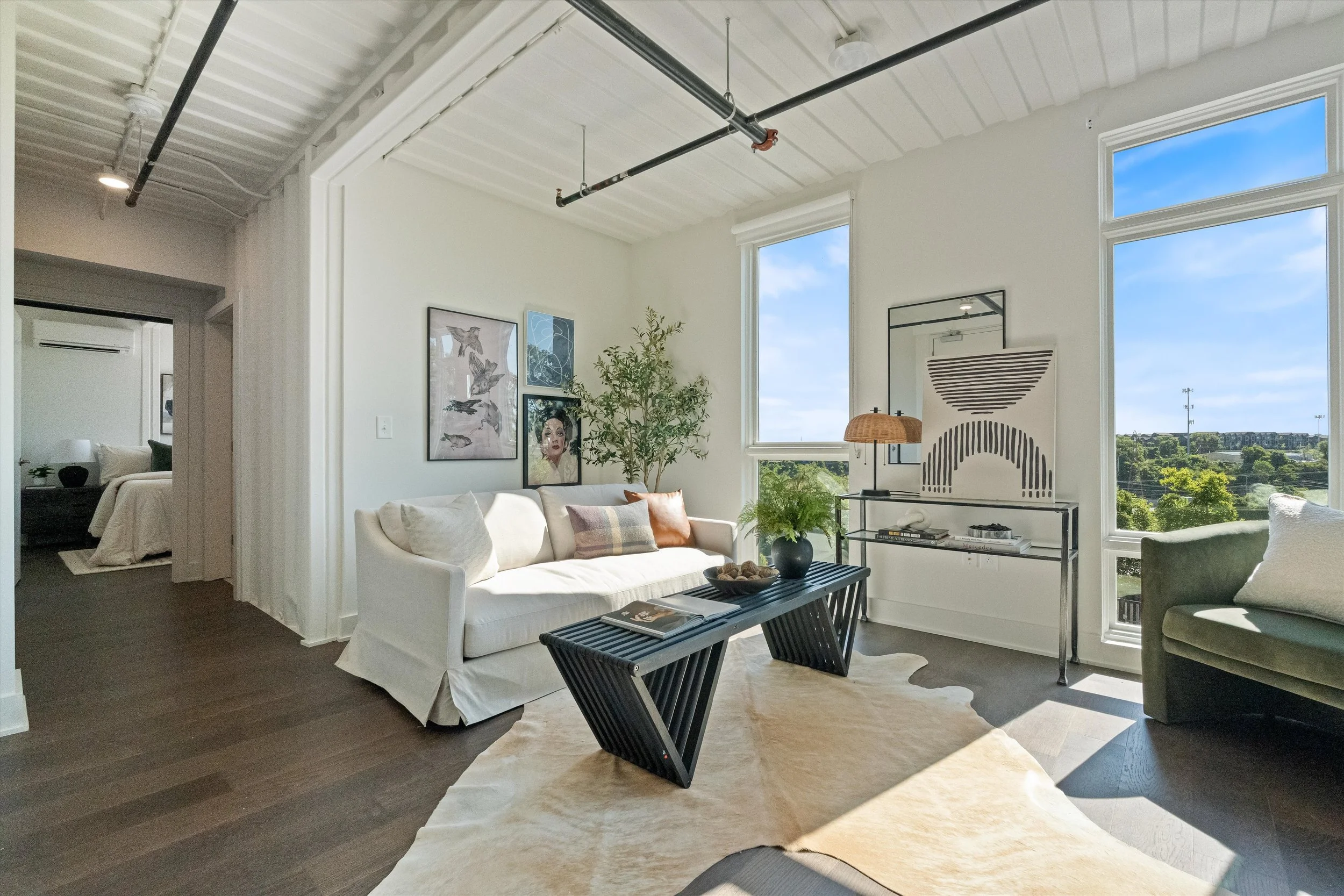 Bright living room with large windows, white sofa with pillows, black coffee table, potted plants, and modern artwork, with a view of a blue sky and green landscape outside.
