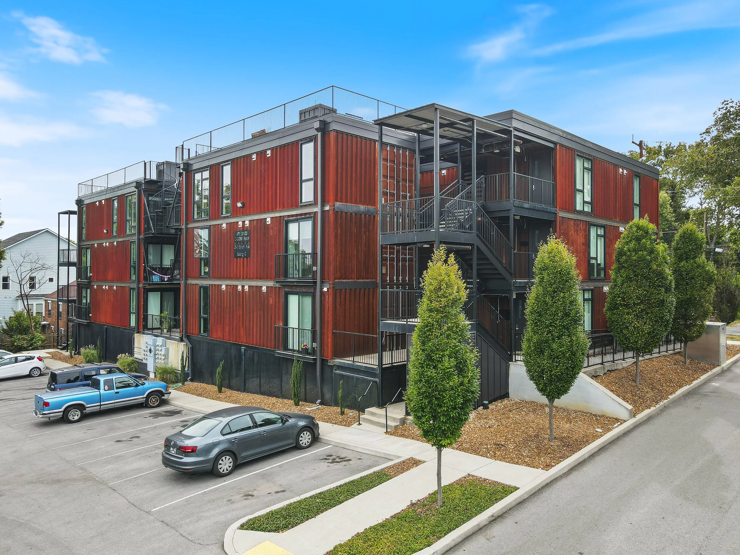 Multi-story apartment building with a red metal exterior, black staircases, and parking lot with a few cars, surrounded by trees and a blue sky.