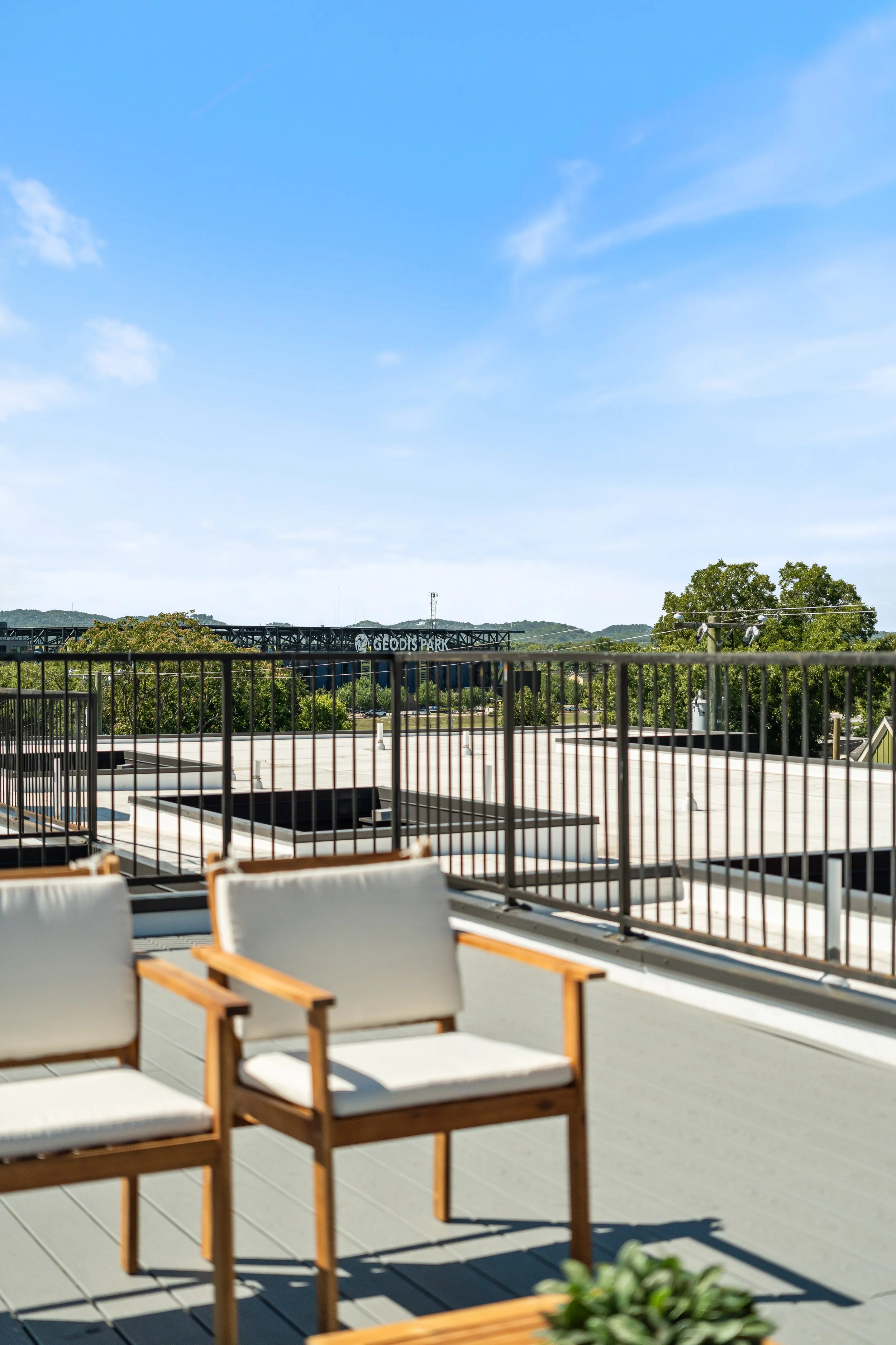 Outdoor rooftop patio with seating, metal railing, and a view of trees and a bridge labeled 'GEODIS PARK' under a blue sky with some clouds.