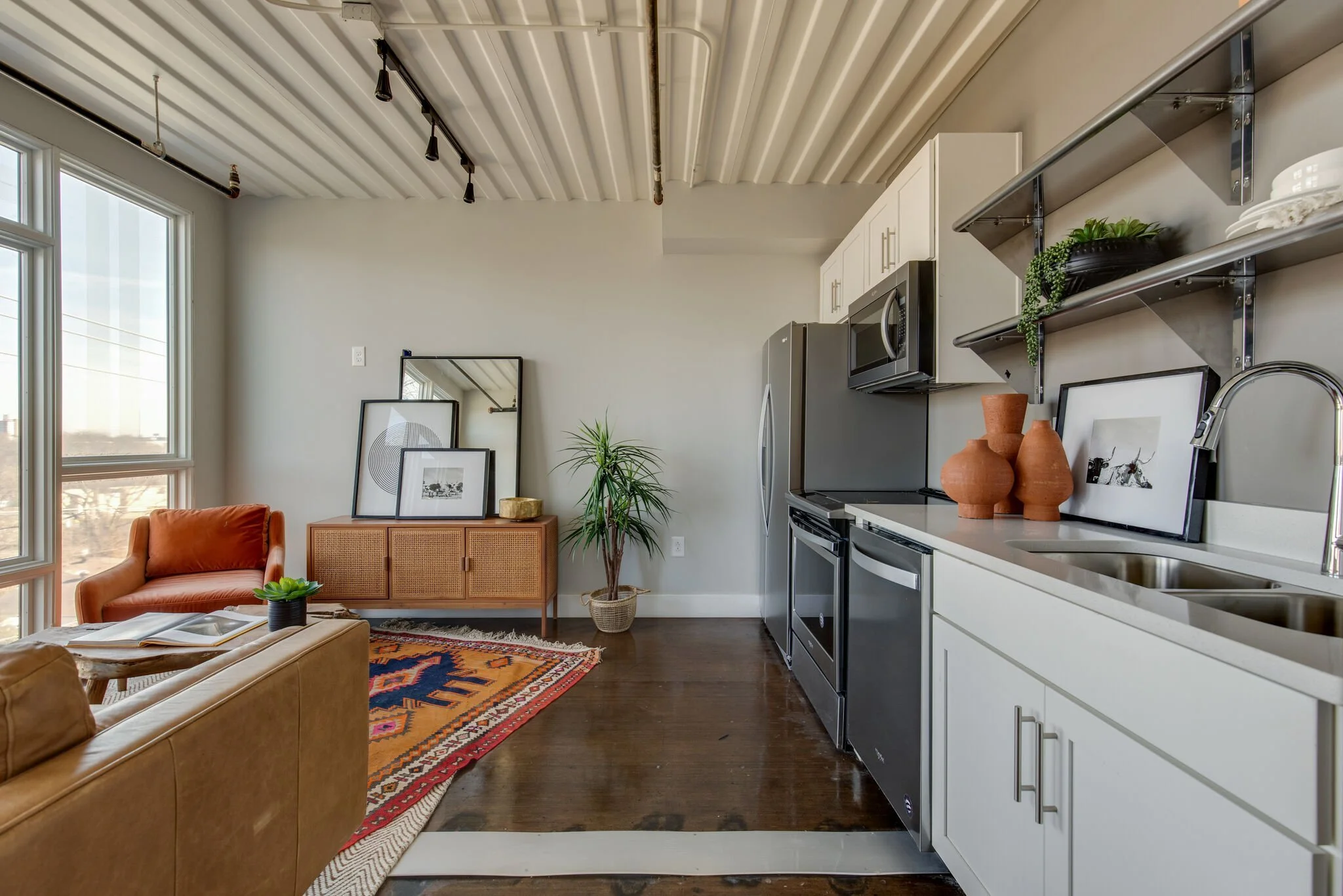 Open-concept living room and kitchen with large windows, white cabinetry, stainless steel appliances, and modern decor, including rugs, framed art, and plants.