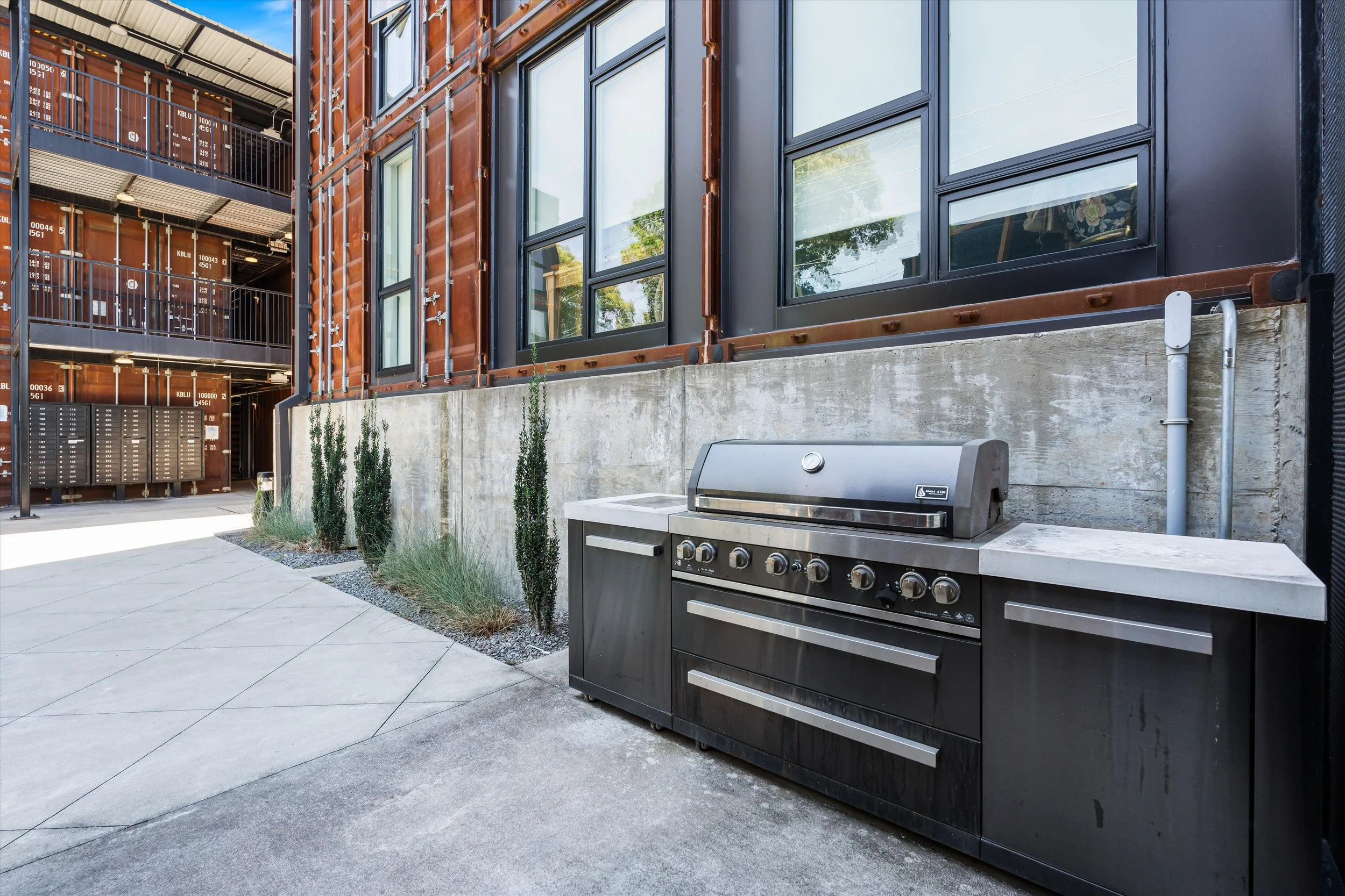 Outdoor patio area with a stainless steel grill and two white countertops, small plants along a concrete wall, and a building with large black-framed windows and a warehouse with shipping containers in the background.