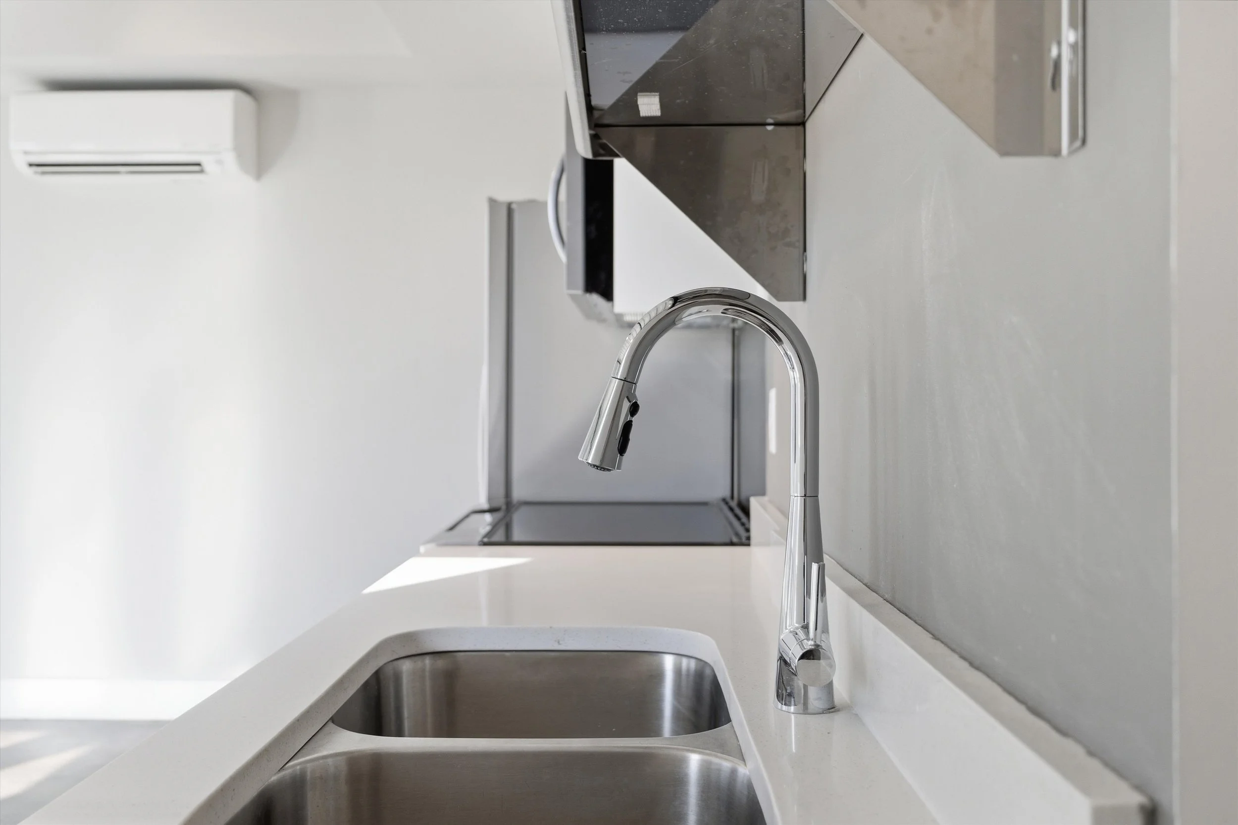 Modern kitchen sink with a chrome faucet and white countertop, with a gray wall and an air conditioning unit in the background.
