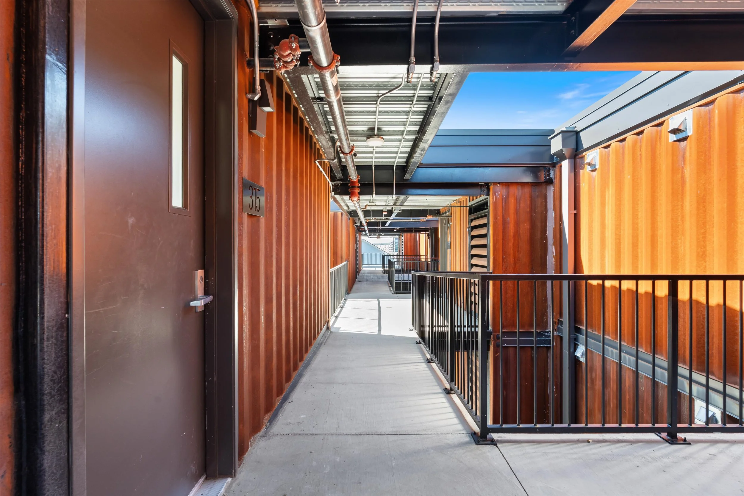 Exterior hallway of a modern apartment building with brown metal doors, black railings, and observe some piping and electrical conduits on the ceiling.