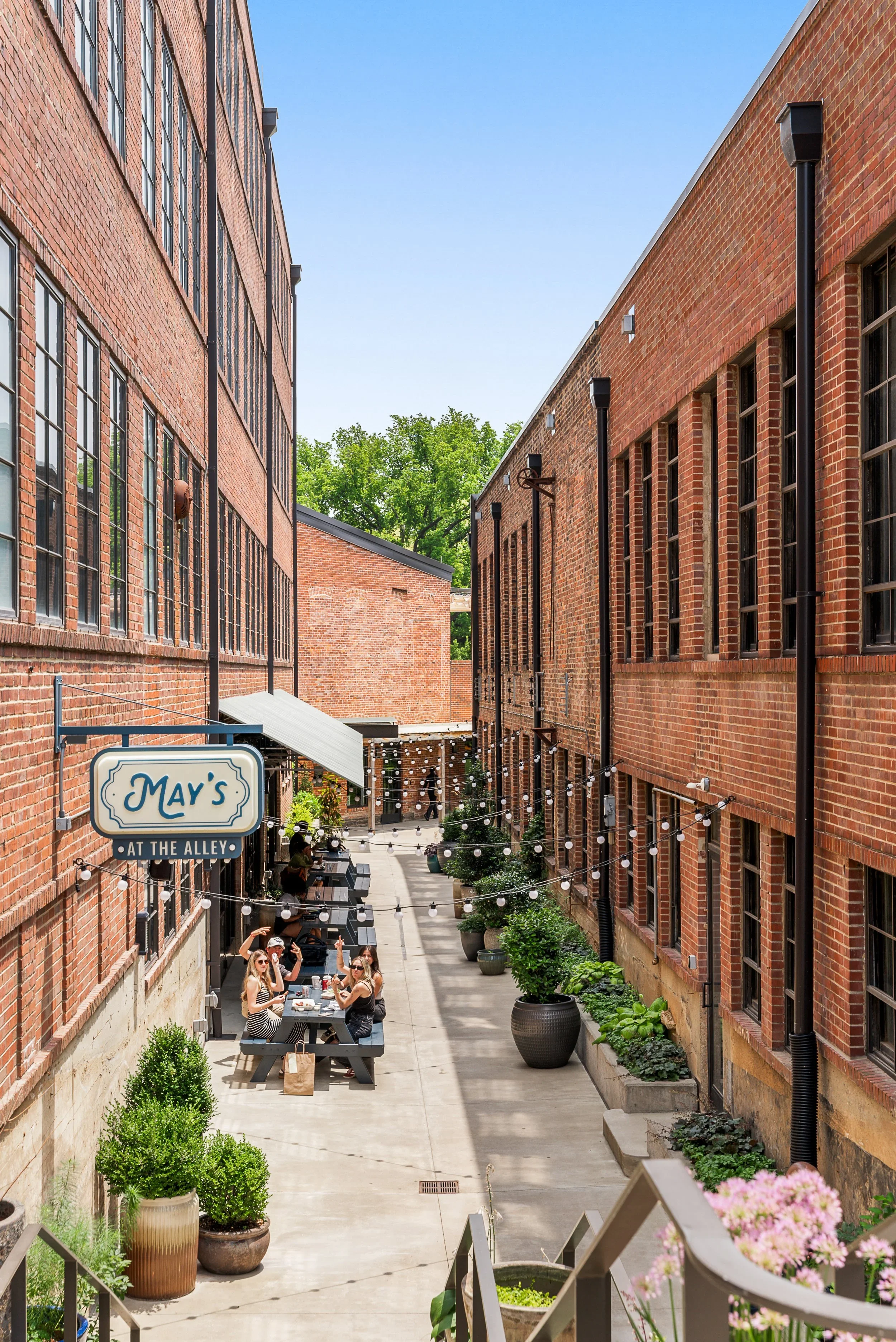 Outdoor patio between two red brick buildings with people dining at tables, string lights, and potted plants. A sign reads 'May's at the Alley'.
