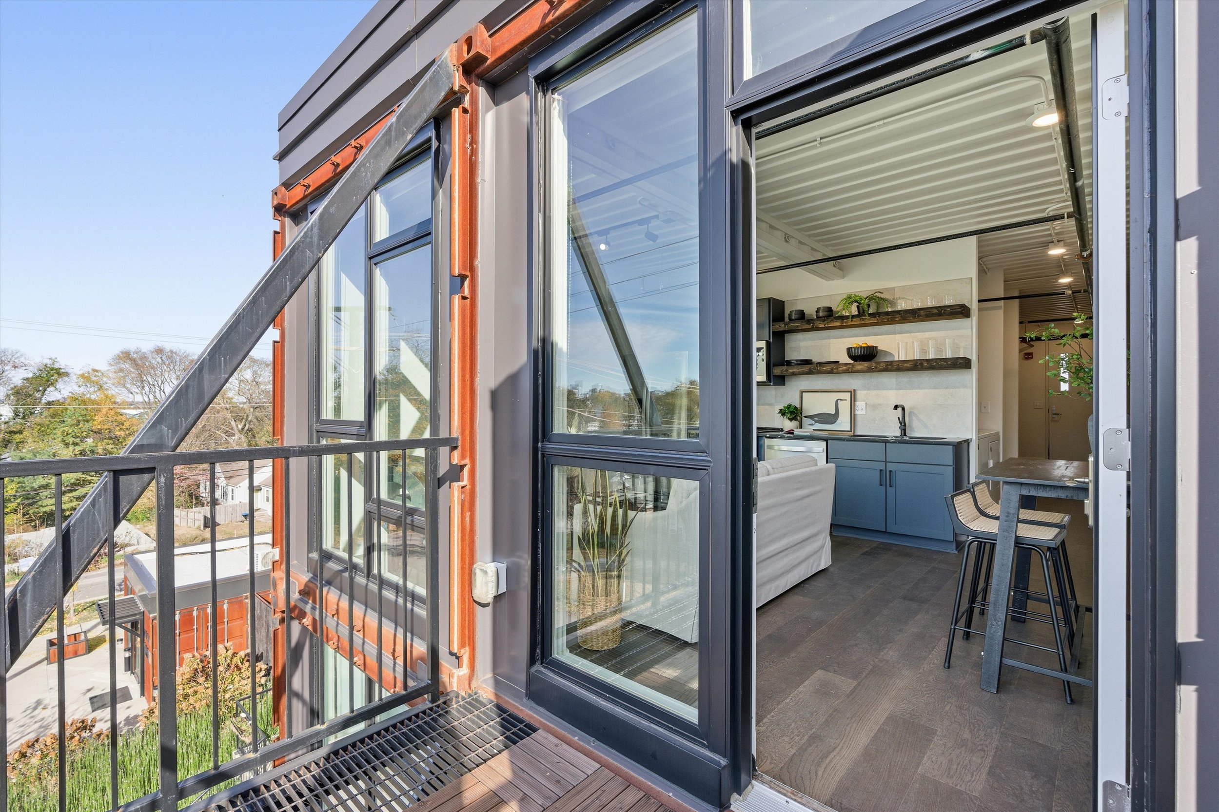 Apartment balcony with black metal railing and wooden floor, leading into a modern kitchen with blue cabinets and open shelves, inside a building with large glass sliding doors, overlooking a neighborhood with trees.