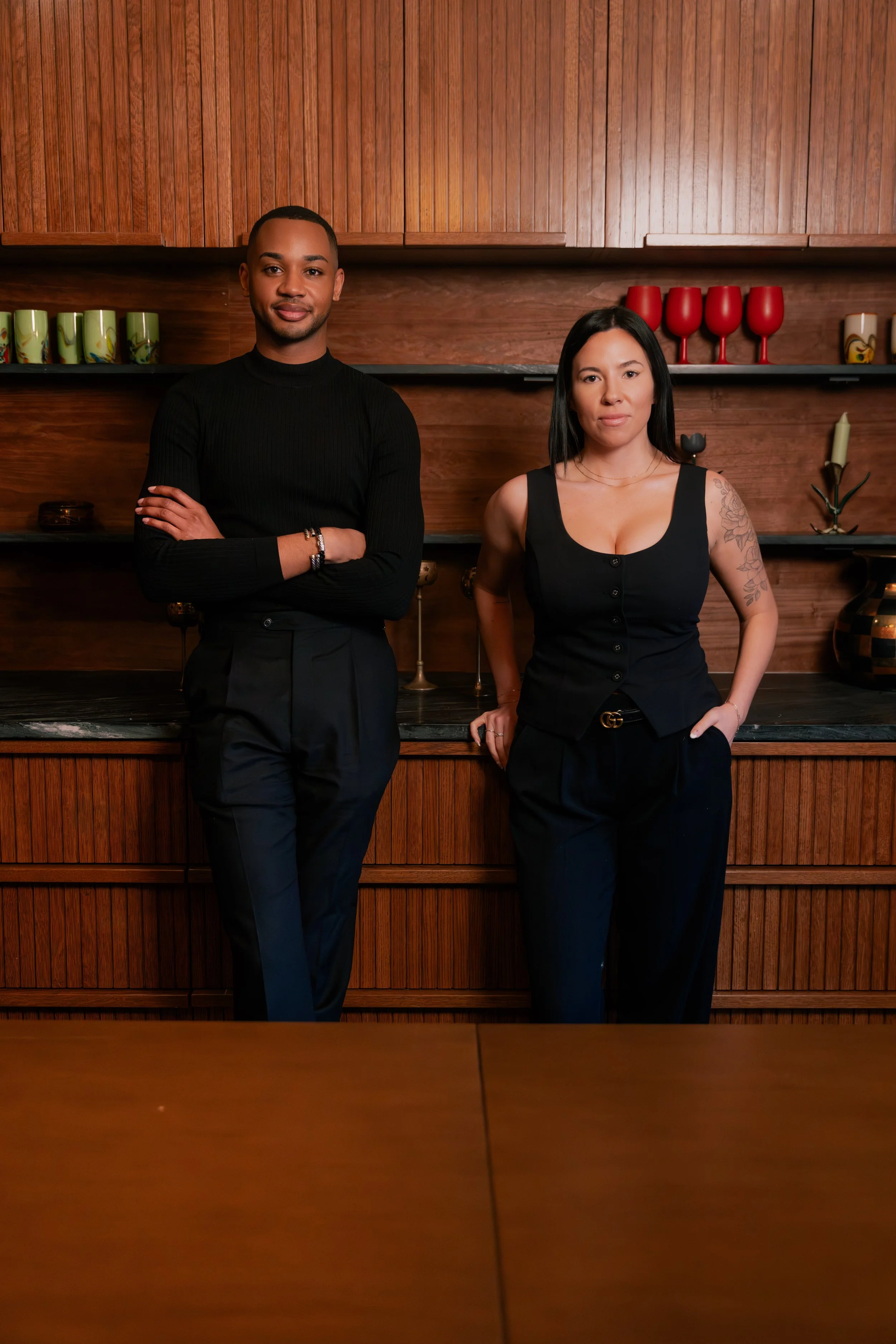 A man and woman standing in front of wooden shelves with decorative items, both dressed in black and looking at the camera.