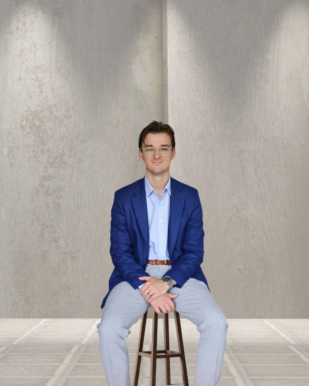A man wearing glasses, a blue blazer, light blue shirt, and gray pants sitting on a wooden stool in front of a plain wall.
