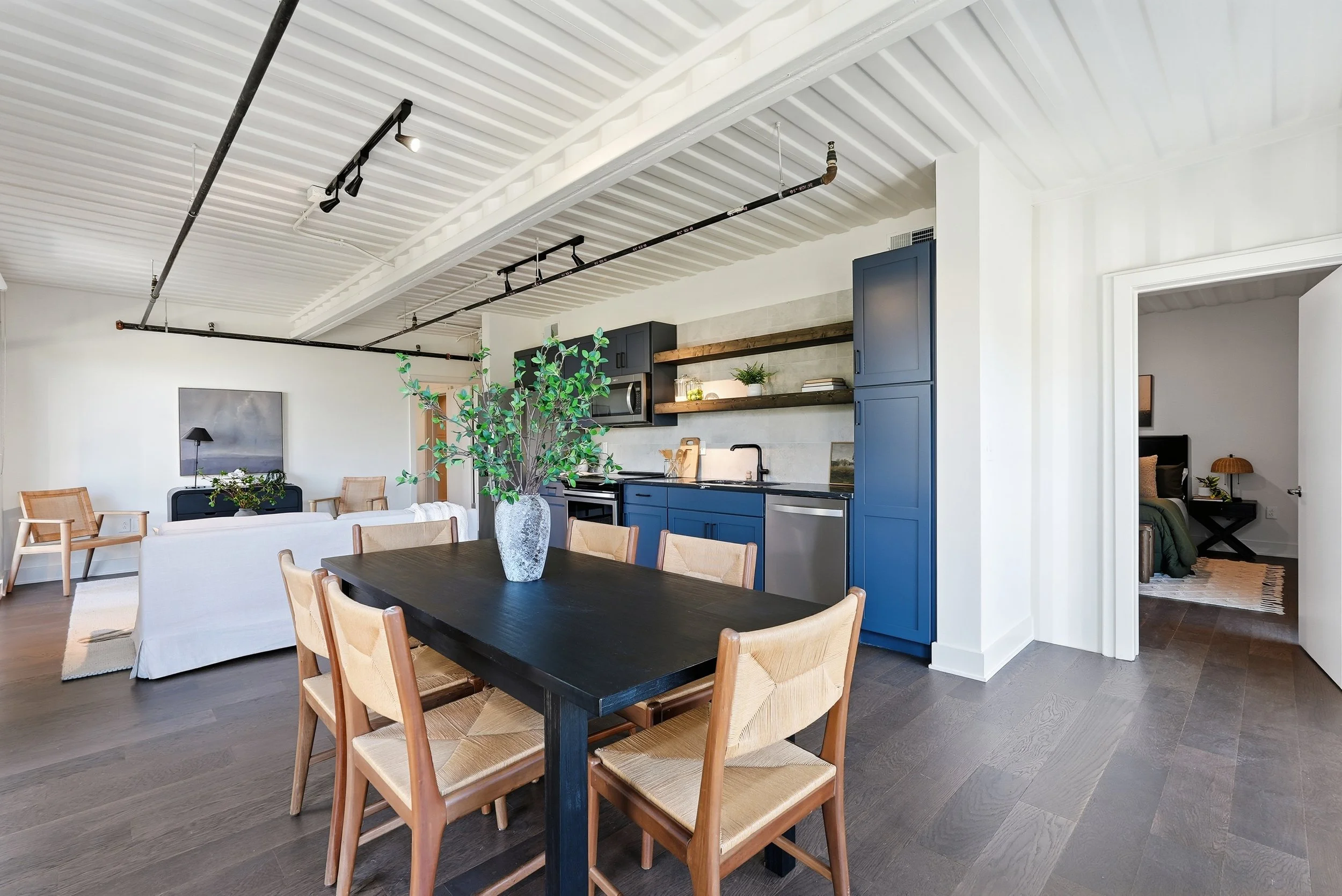 Open-concept living room and kitchen with blue cabinets, a dining table with wooden chairs, and a bedroom visible through a door.