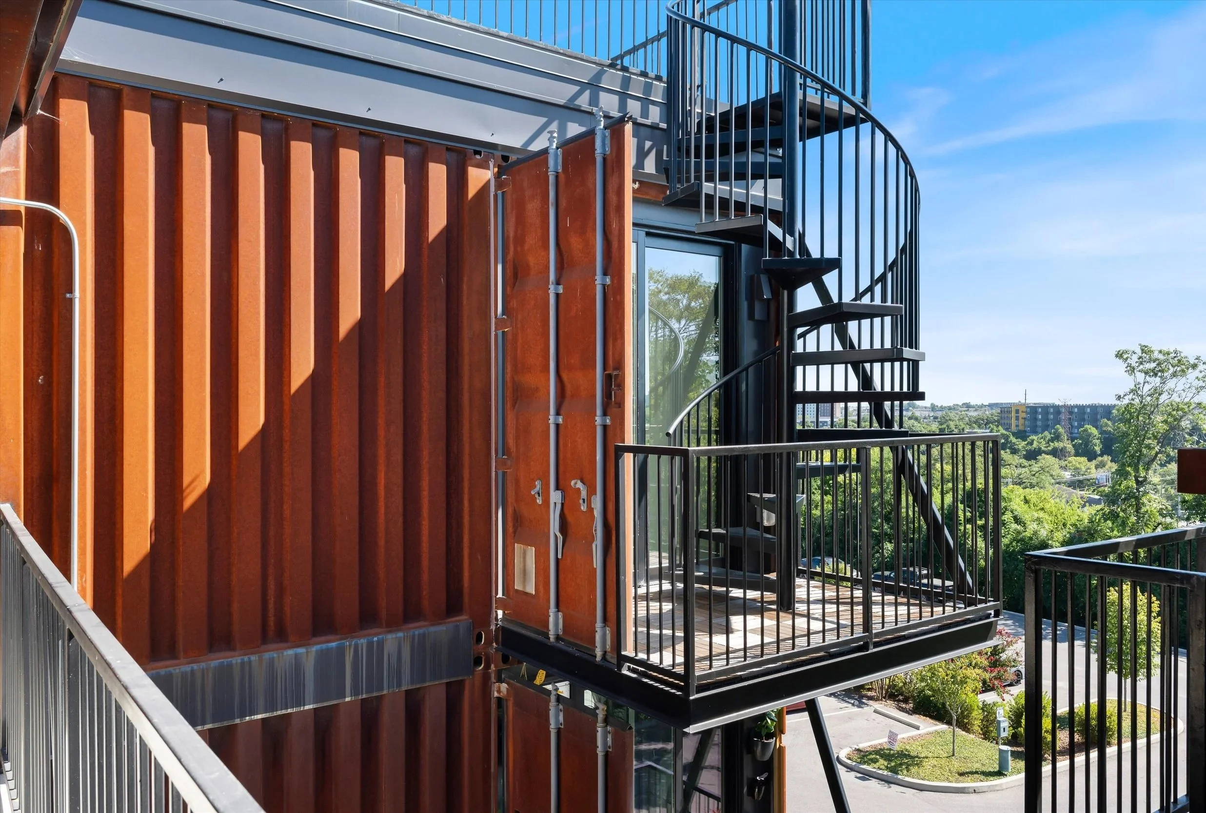 Exterior view of a modern building with a black spiral staircase and small balcony on the upper floor, metal railing, and surrounding greenery.