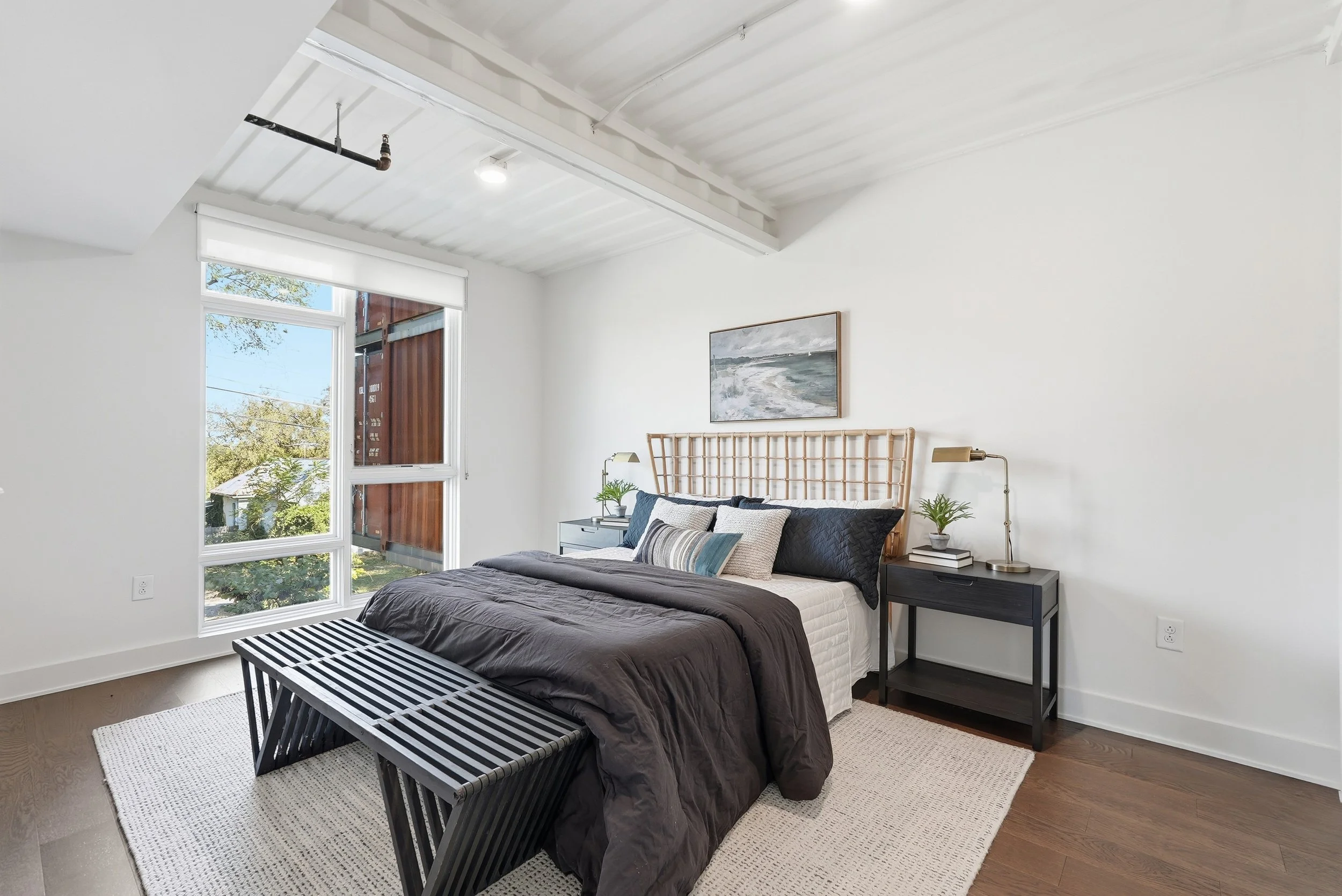 Modern bedroom with white walls, large window, bed with black bedding and various pillows, two black nightstands with lamps and plants, landscape painting above the bed, wooden bench at the foot of the bed, beige rug, hardwood floors, ceiling with ex