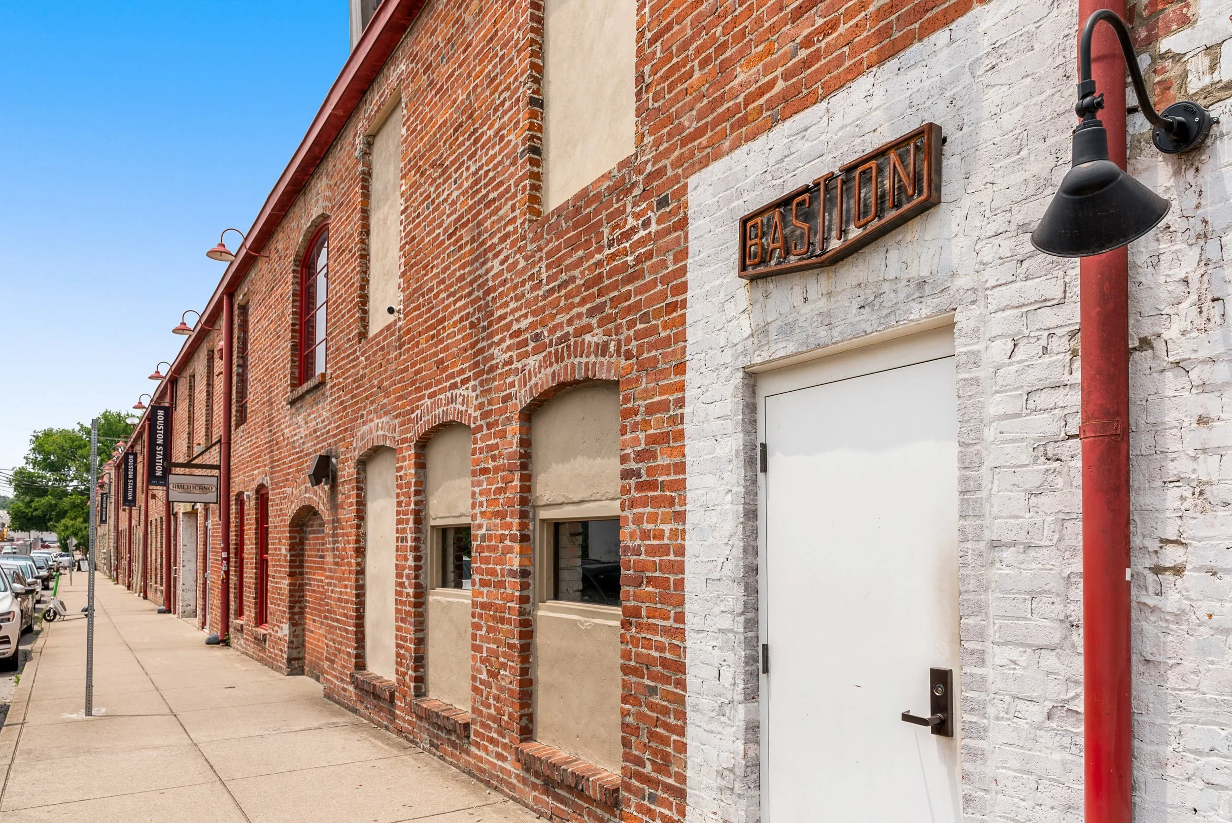 Brick building on a sidewalk with multiple lit signs, white door, and a black light fixture, with a cloudy blue sky in the background.