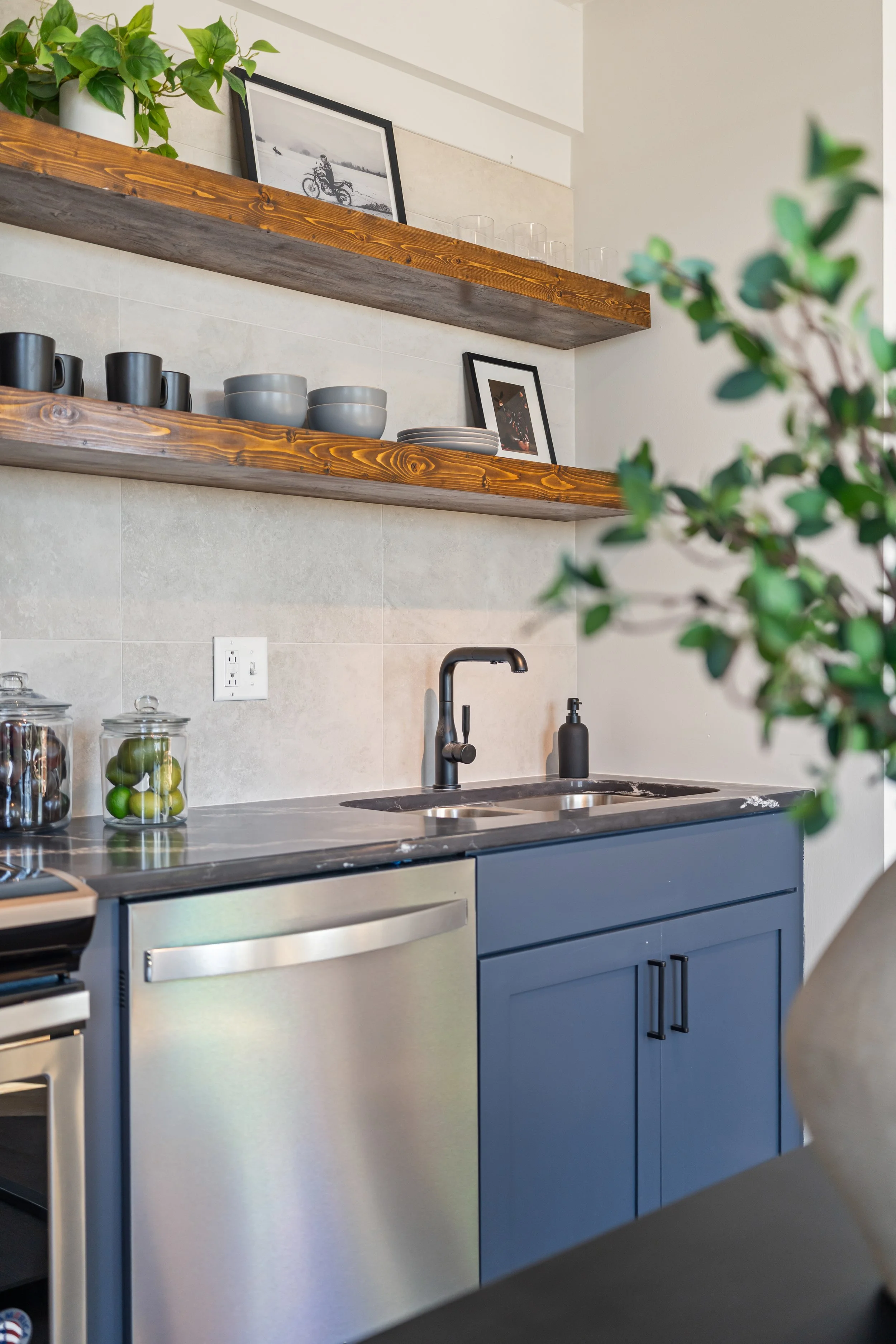 Modern kitchen with blue cabinets, black faucet, open wooden shelves with picture frames, cups, bowls, and glass jars, and a potted plant.