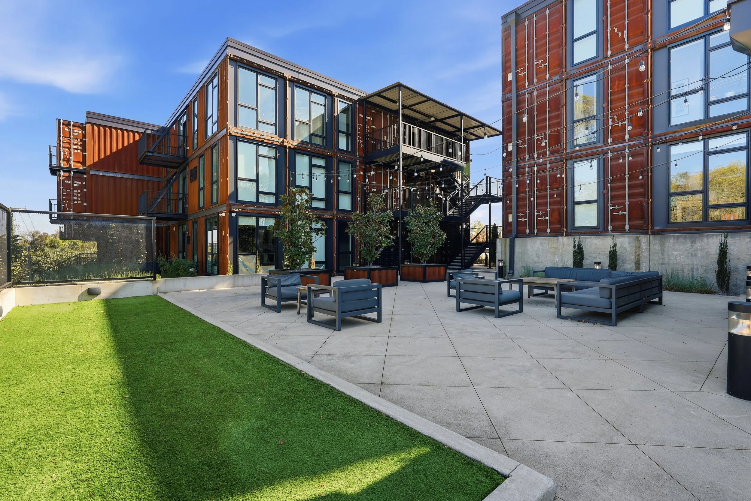 Outdoor patio with modern seating area, adjacent to a building made of repurposed shipping containers, featuring large glass windows, string lights, and potted trees, under a clear blue sky.