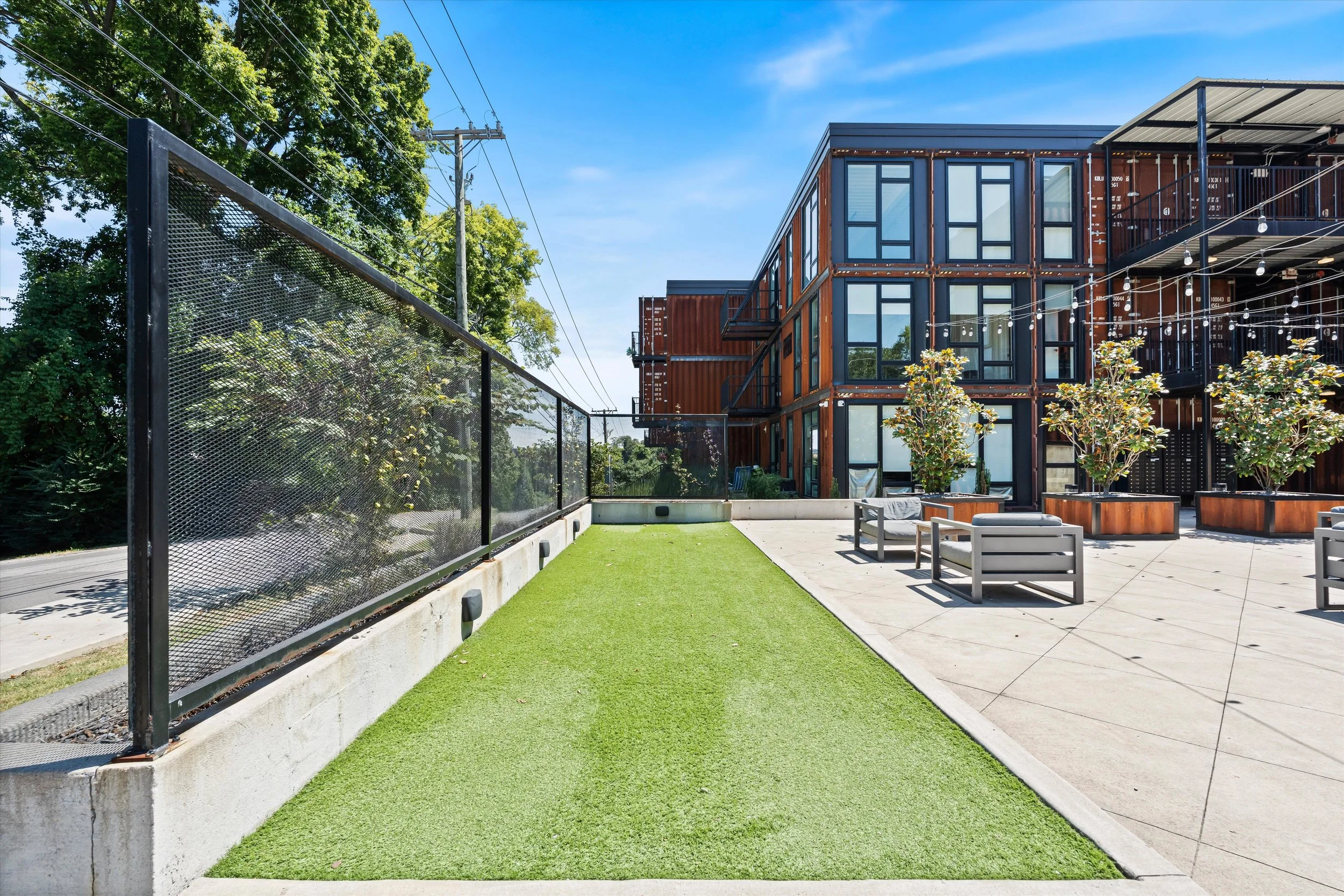Modern apartment building with container-style design, large windows, outdoor seating, planter boxes with trees, string lights, green artificial grass area, and a black metal fence on the left, under a clear blue sky.