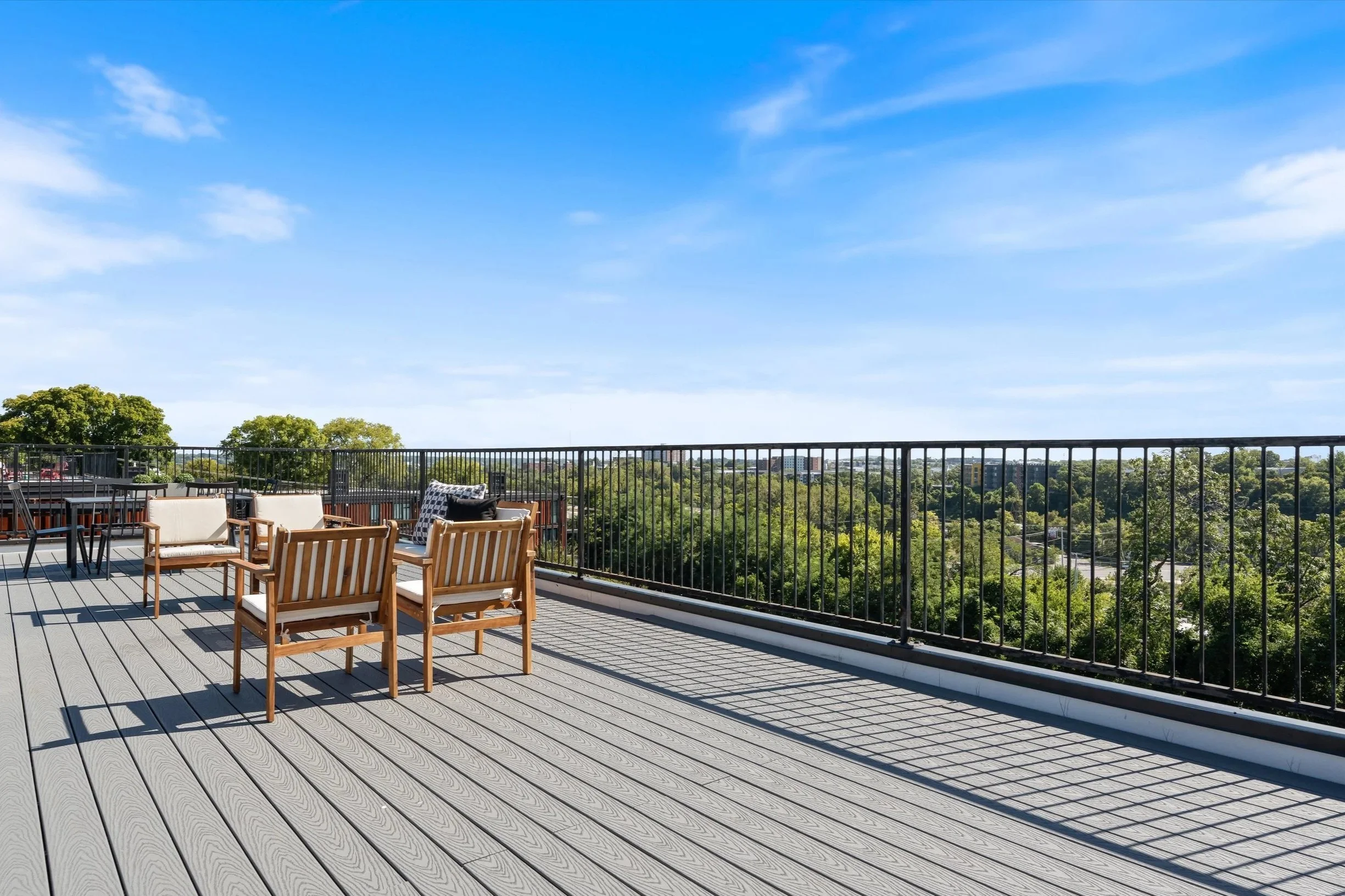 Rooftop terrace with wooden and cushioned seating, overlooking a city with green trees and a blue sky with a few clouds.