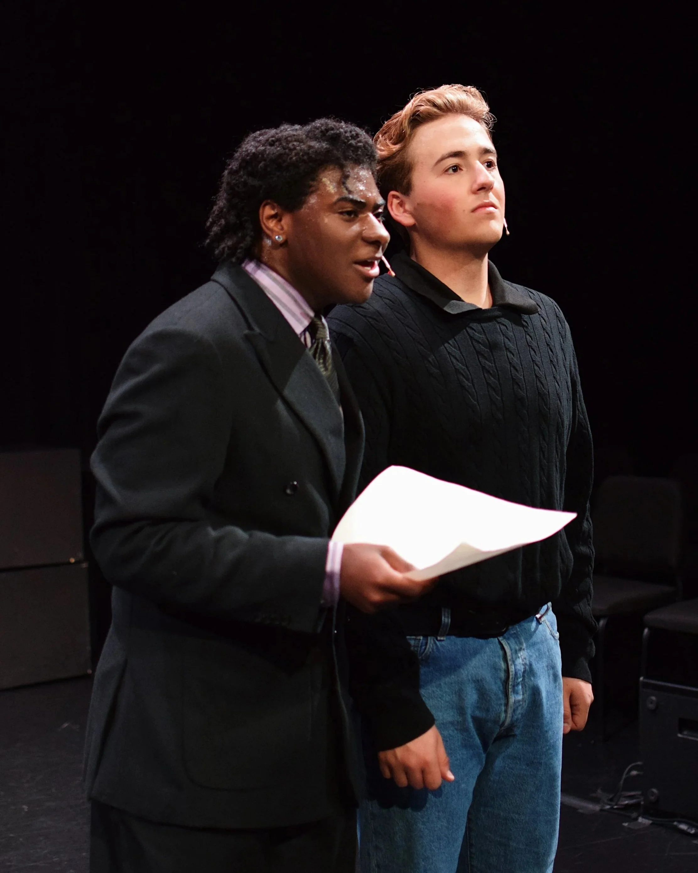 Two young men standing on a dark stage, wearing casual and semi-formal clothing, with one holding a piece of paper and the other looking forward. They appear to be participating in a theatrical or presentation event.