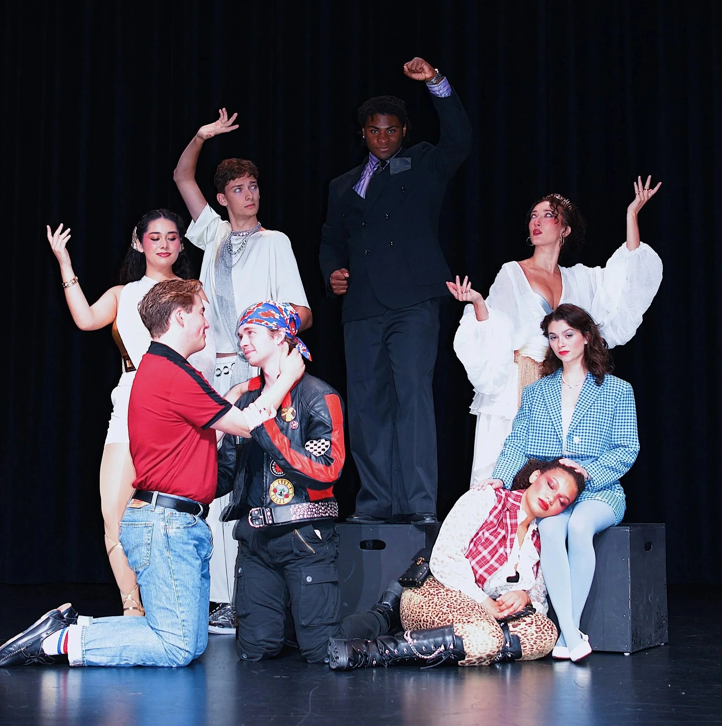 The image shows a group of young people on stage, striking various poses, with some kneeling and others standing, against a black curtain background, possibly performing or rehearsing.