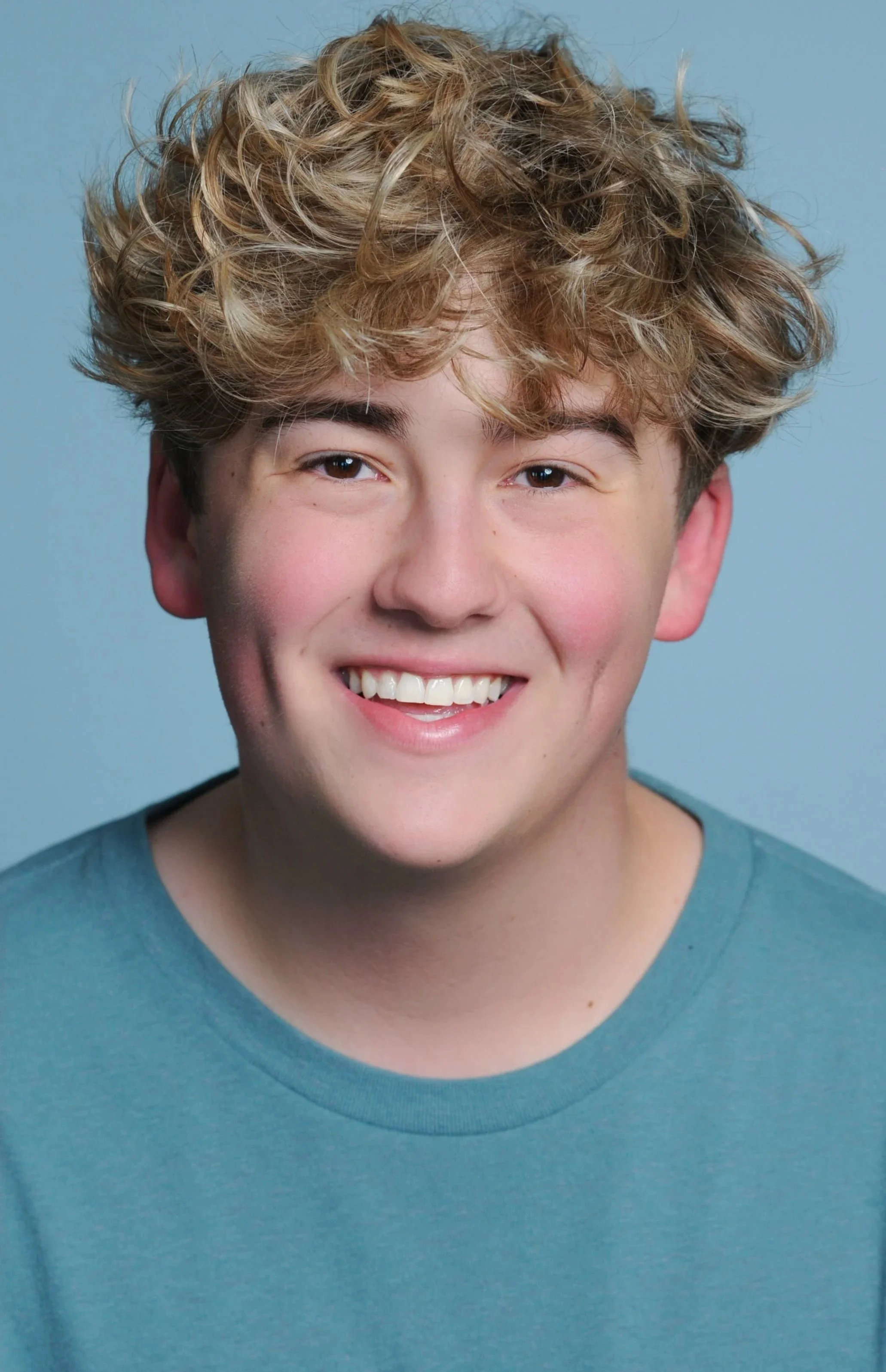 A smiling young man with curly blonde hair wearing a blue shirt against a light blue background.