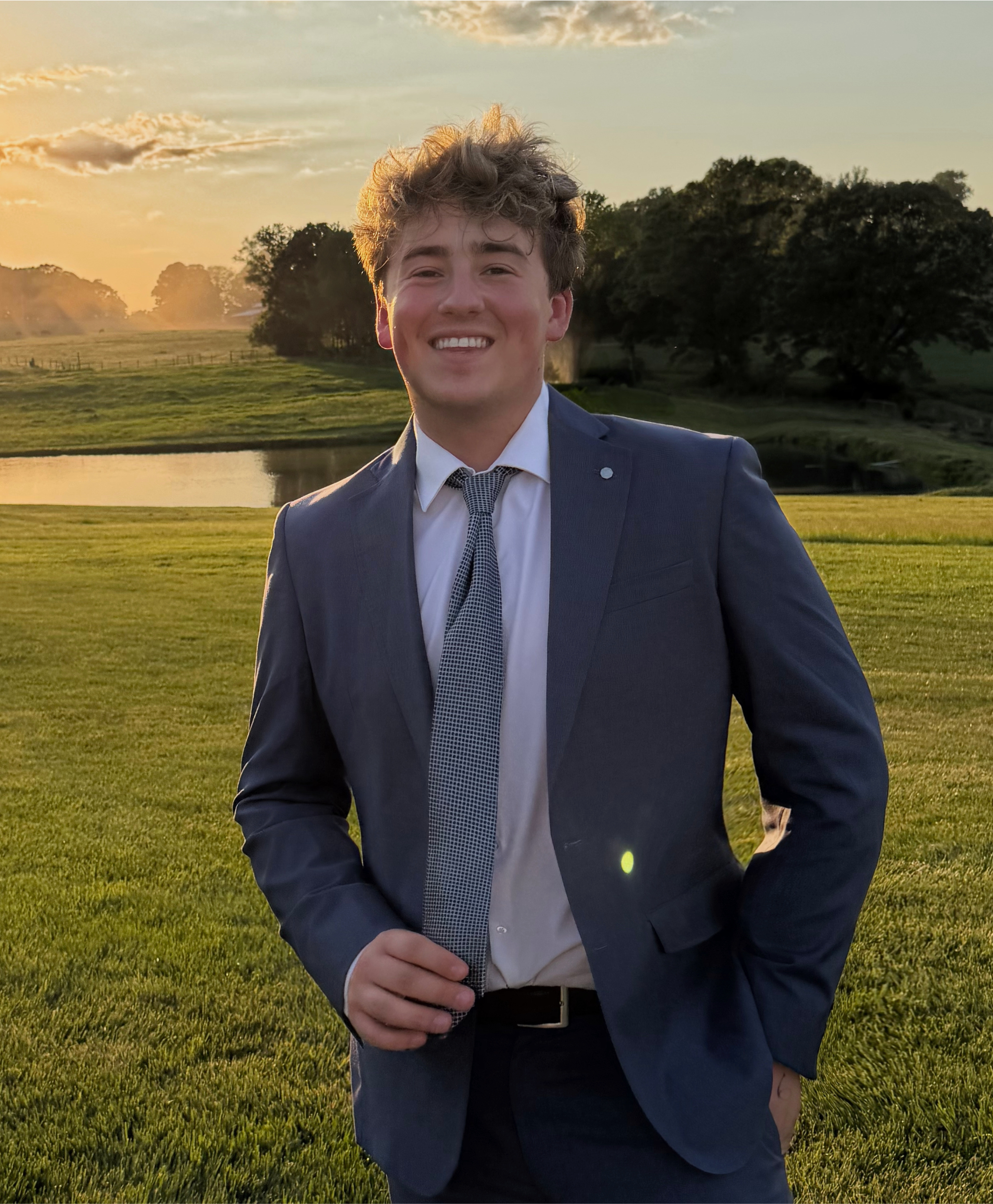 A young man smiling and dressed in a business suit, standing outdoors during sunset with green grass, a pond, and trees in the background.