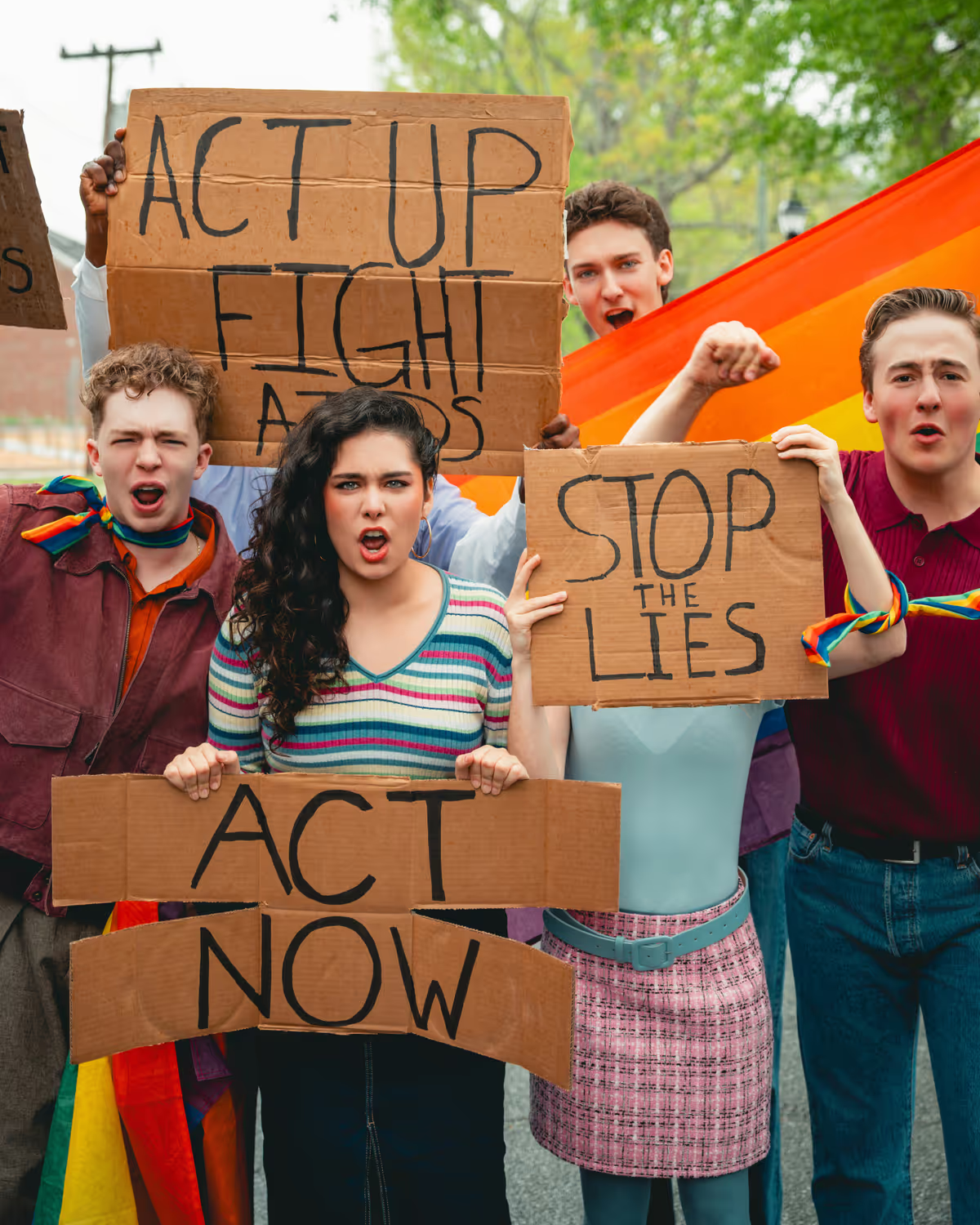 Group of diverse young people protesting for LGBTQ+ rights outdoors, holding signs that read "ACT UP FIGHT AIDS," "STOP THE LIES," and "ACT NOW," with rainbow flags and rainbow-colored accessories.