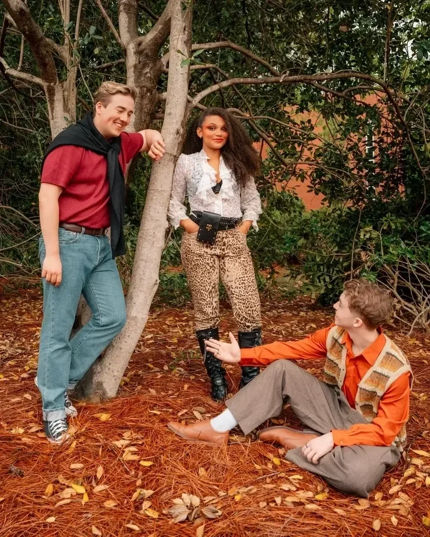 Three people outdoors in a wooded area with fallen leaves on the ground. One person is sitting on the ground wearing an orange shirt and patterned vest, reaching out to another person standing near a tree, wearing a white ruffled shirt and animal print pants. A third person, leaning against a tree, is smiling, wearing a red shirt with a black sweater tied around their shoulders.