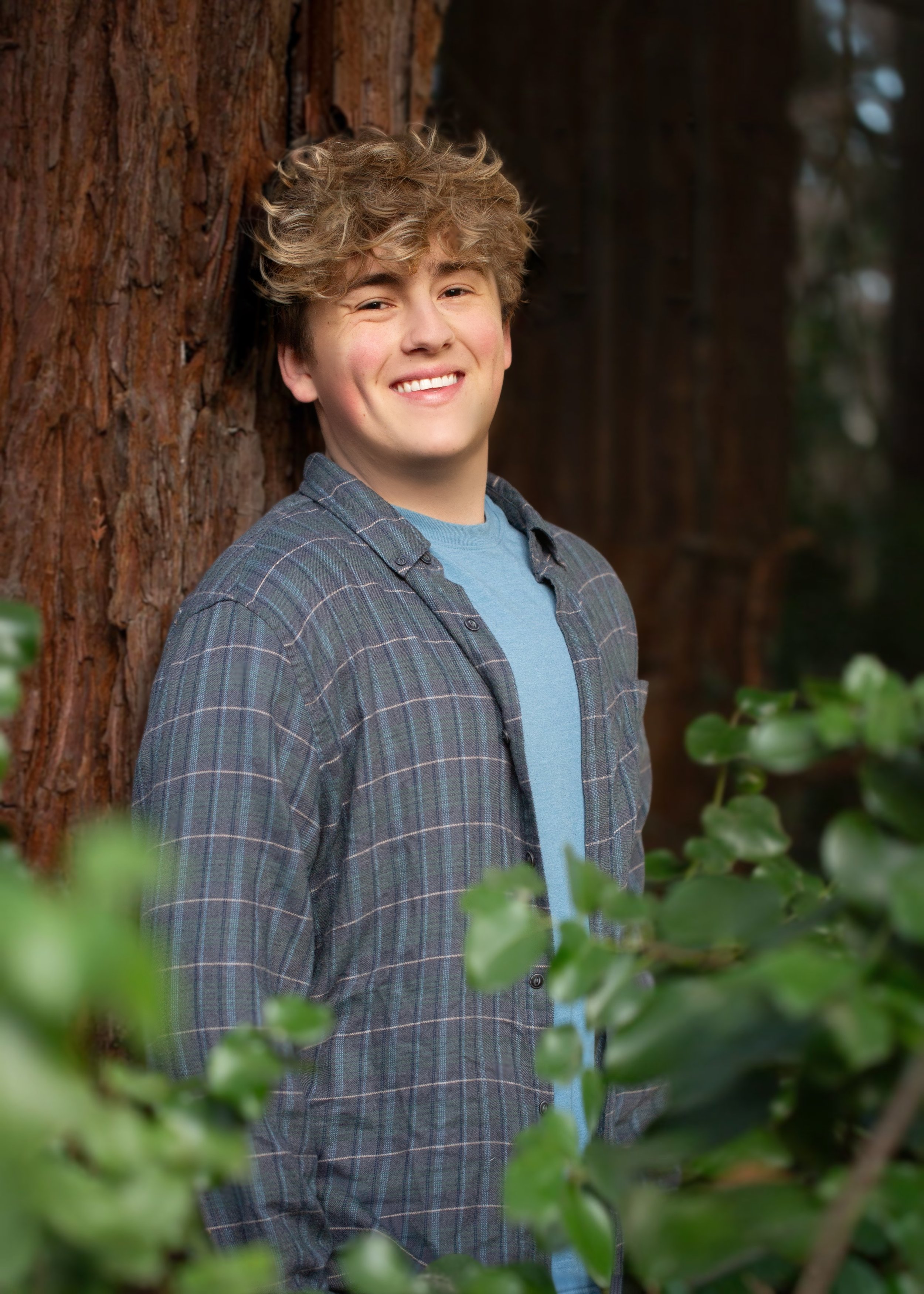 A young man with curly blond hair, smiling, standing outdoors next to a large tree and surrounded by green foliage.
