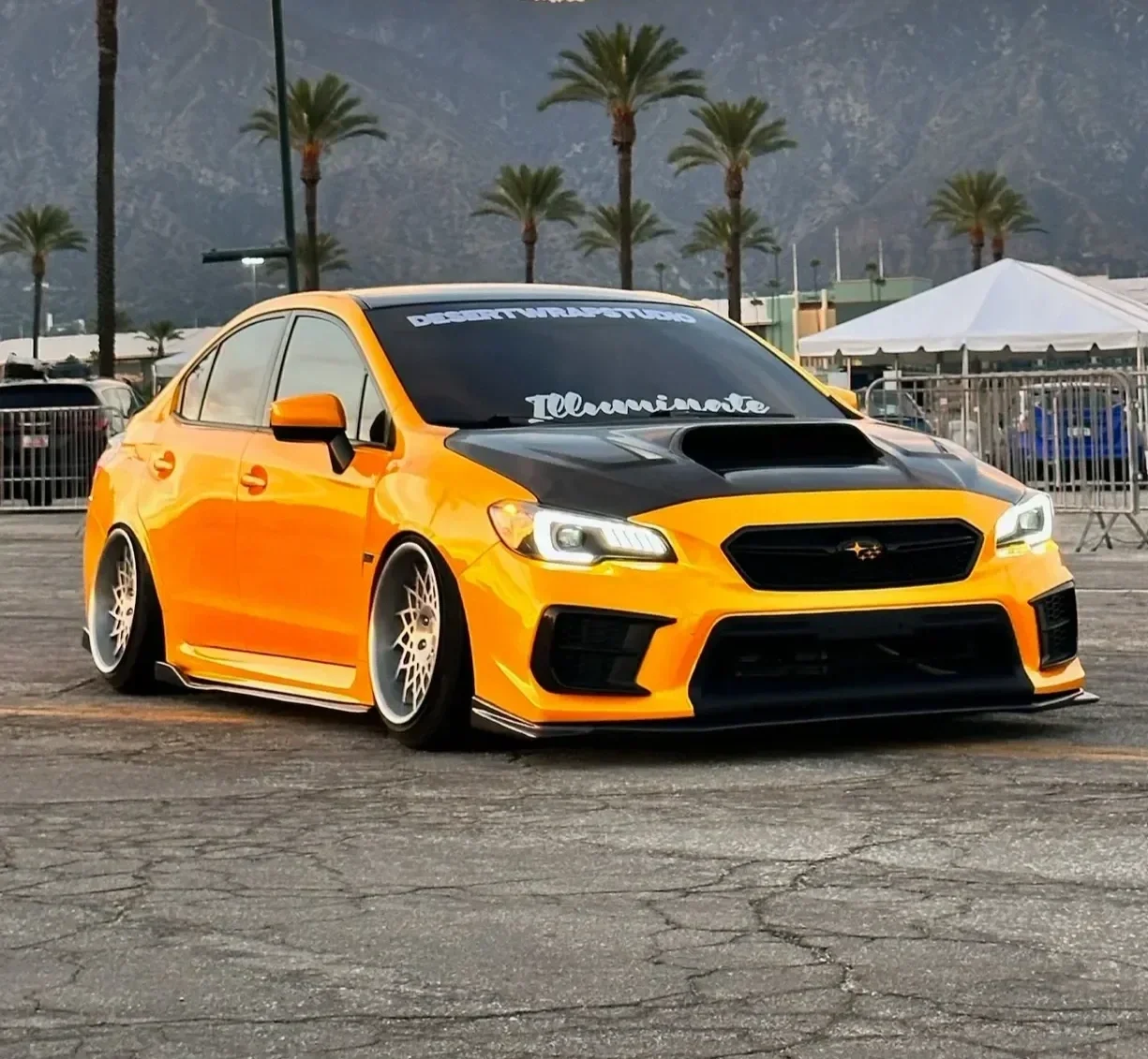 A yellow Subaru sedan with black accents, lowered suspension, wide wheels, and modified body kit on a parking lot with palm trees and mountains in the background.