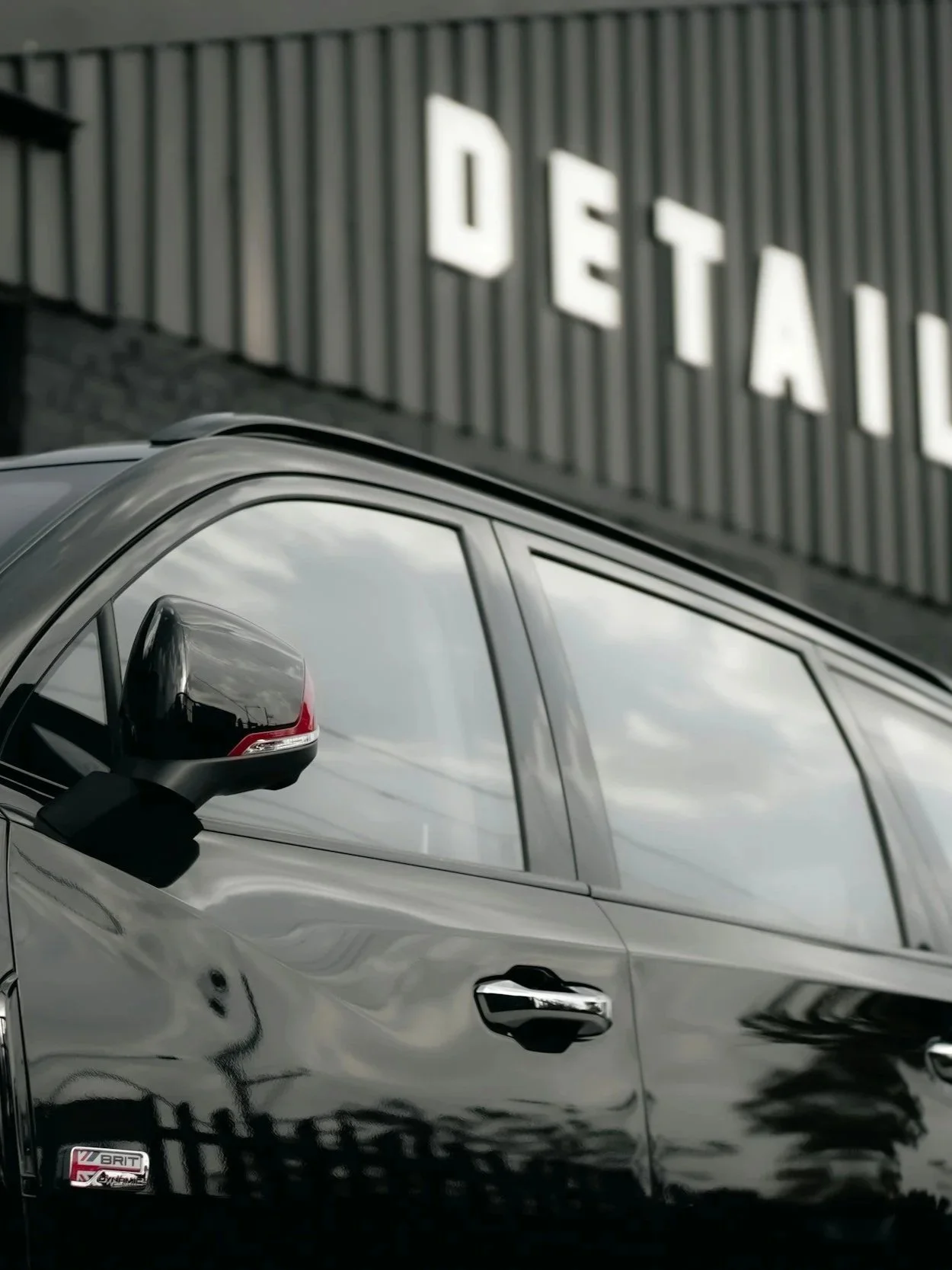 Close-up of a silver luxury SUV parked outside a building with a sign that says 'DETAI' in large white letters on a grey corrugated metal wall.