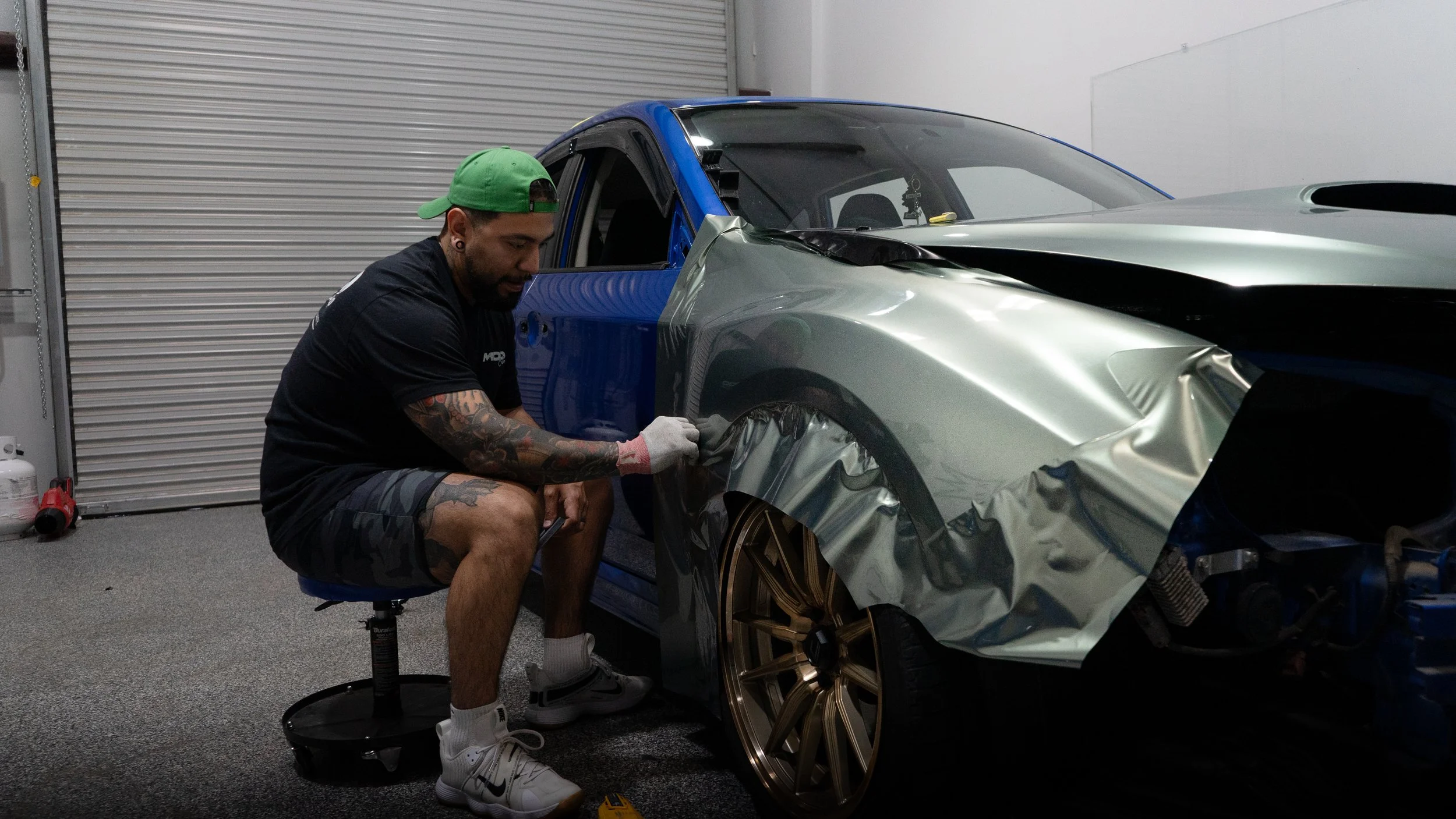 A man working on a car in a garage, applying protective film to the front fender. The car is partially disassembled, with the front bumper removed, revealing the inner components. The man is sitting on a stool, wearing a black t-shirt, camouflage shorts, white sneakers, a green cap, and has tattoos on his arms.