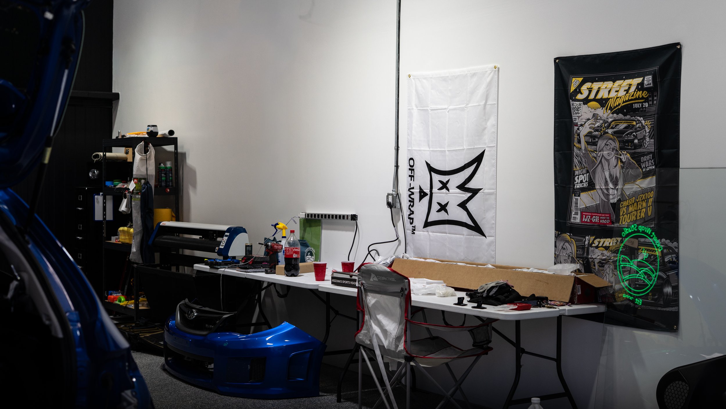Workshop with car parts, tools, and posters on the wall, including a large blue car bumper on the table and a racing-themed poster.