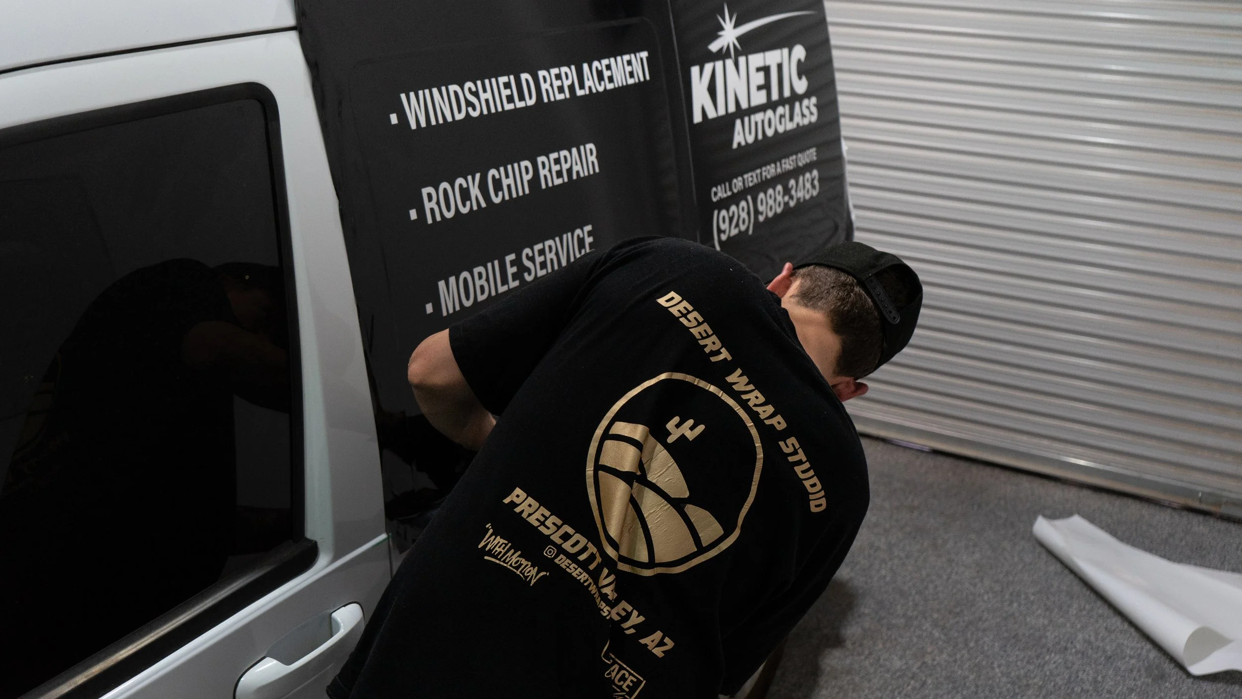 A person installing tinted film on a car window in a garage. The garage door is metal, and the person is wearing a black hoodie with a logo on the back.