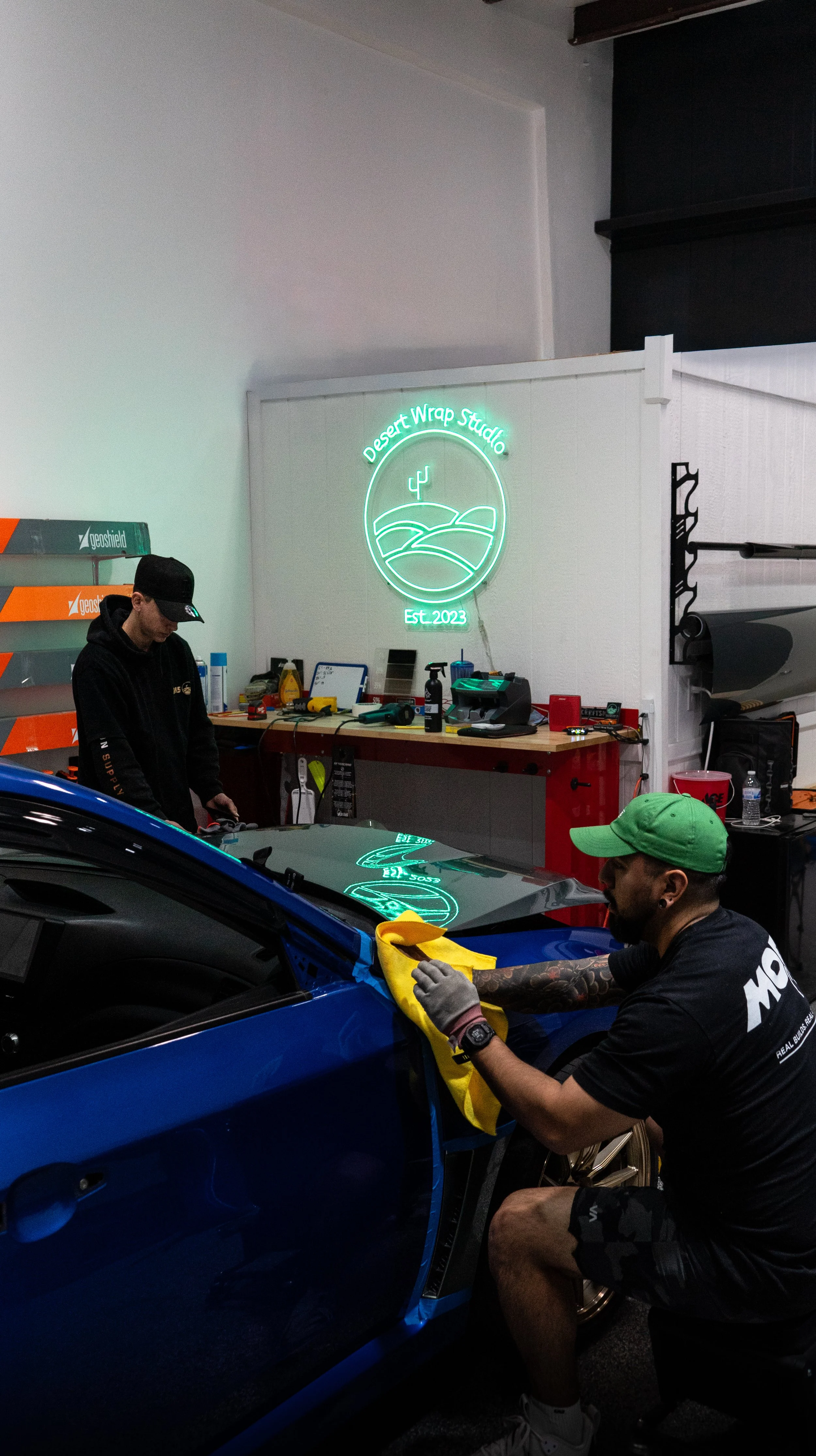 Two men working on a blue car inside a garage, with a neon sign that reads 'Desert Wrap Studio' and 'Est. 2023' on the wall behind them.