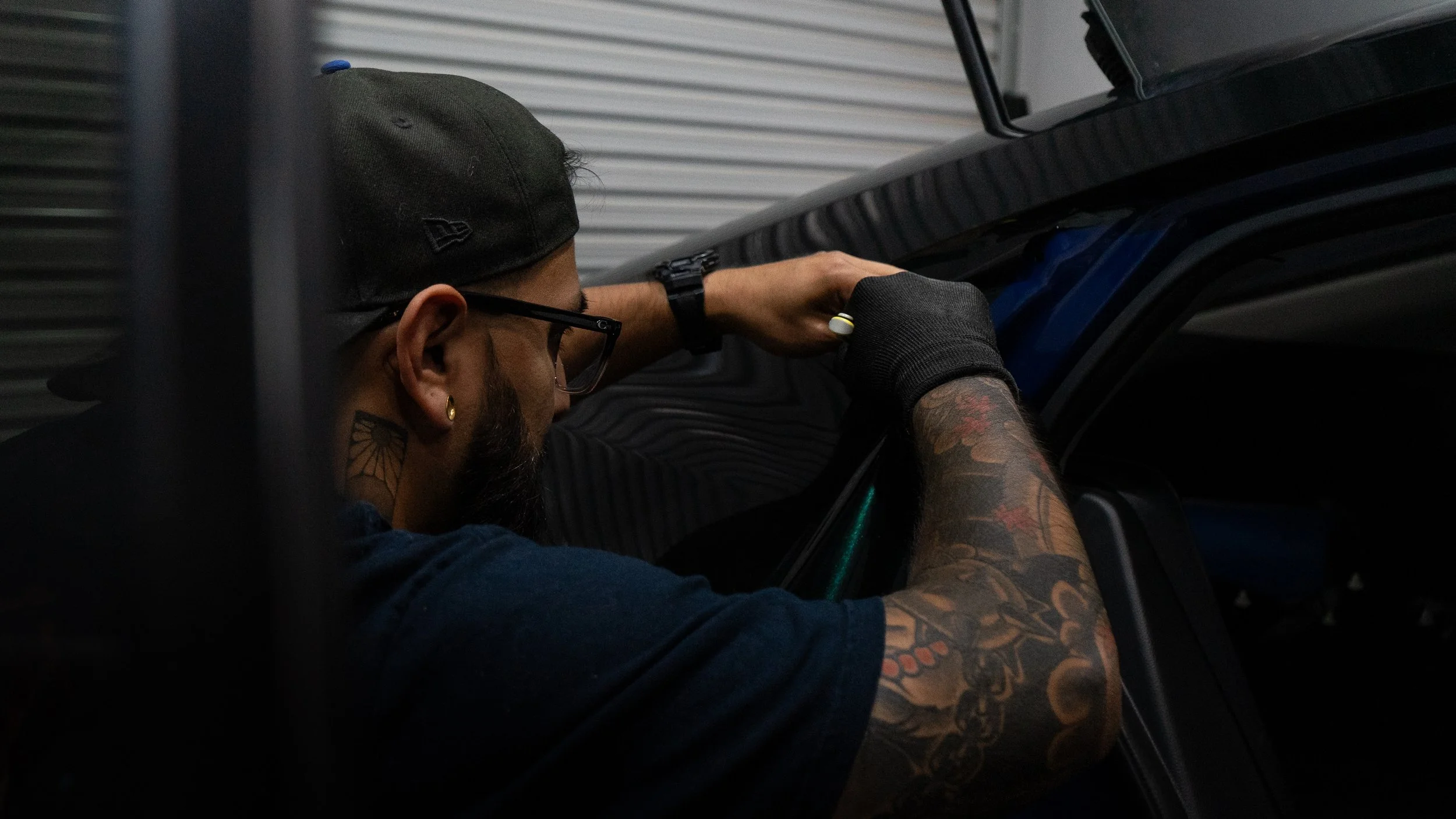 A man with tattoos on his arm, wearing a baseball cap and glasses, working on a vehicle in a garage with metal walls.